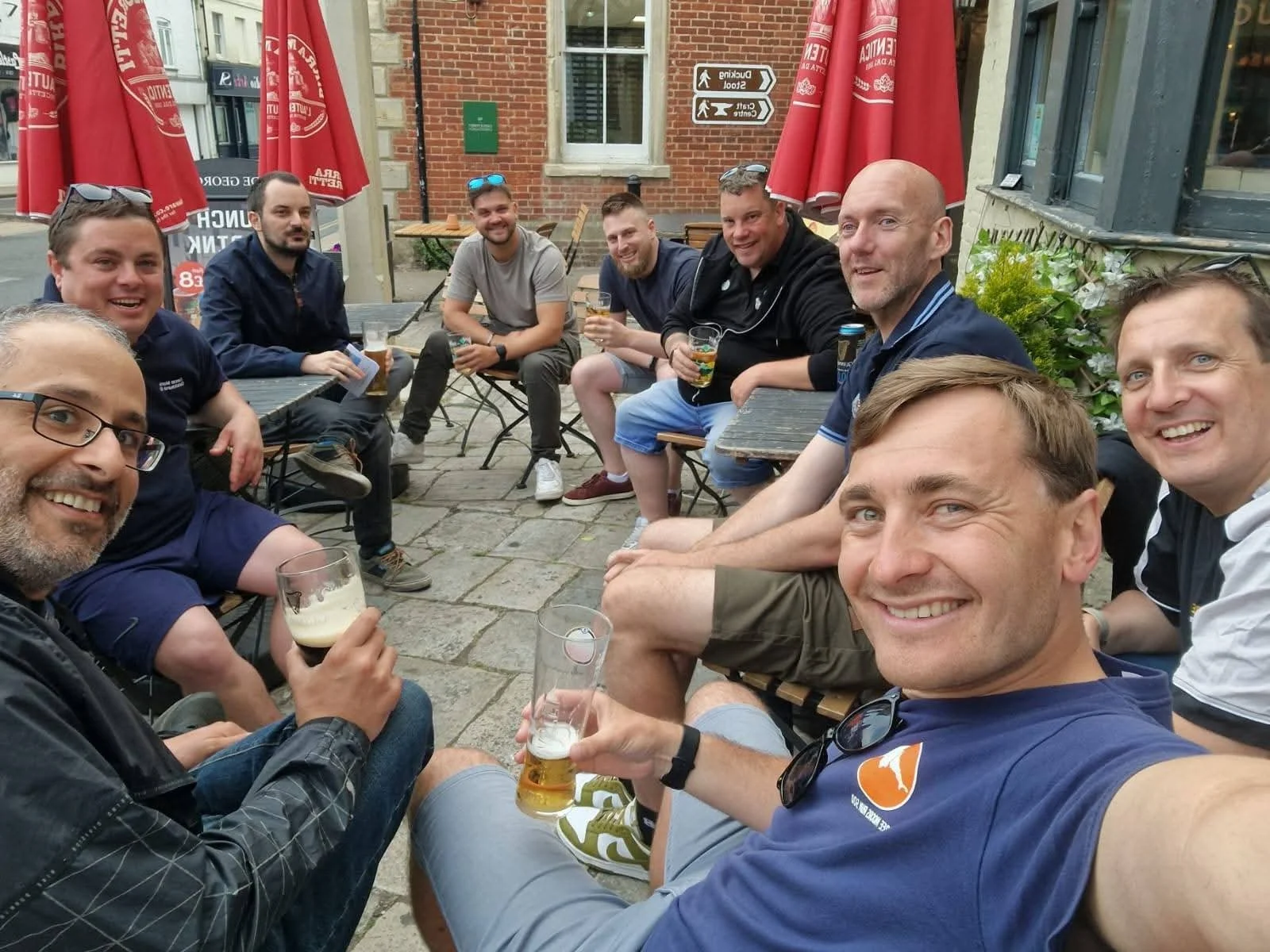 Group of men sitting around outdoor table at a pub, smiling and holding drinks.