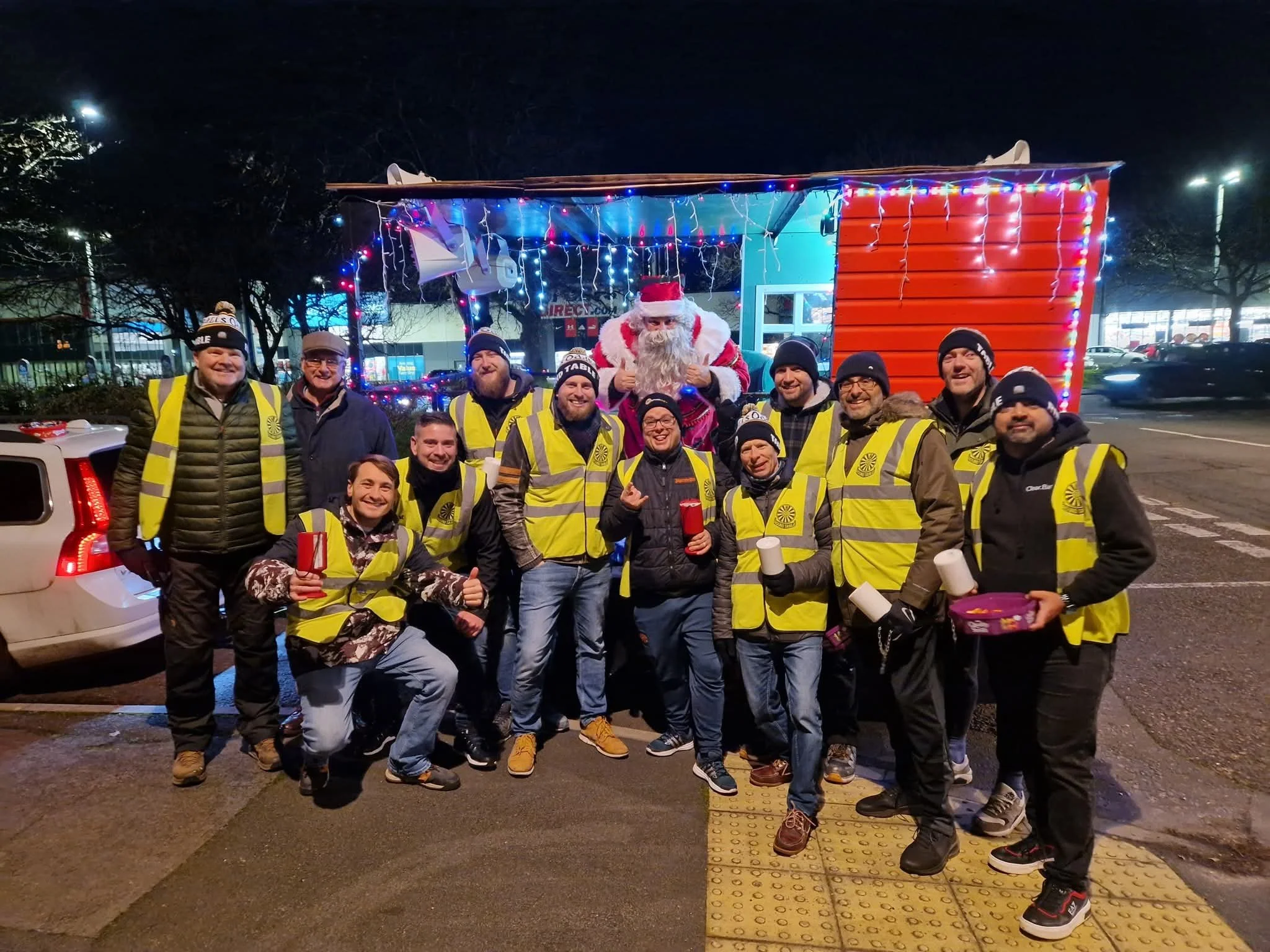Group of people wearing yellow safety vests gathered at night around a Santa Claus figure in a Christmas-themed setup with Christmas lights and decorations.