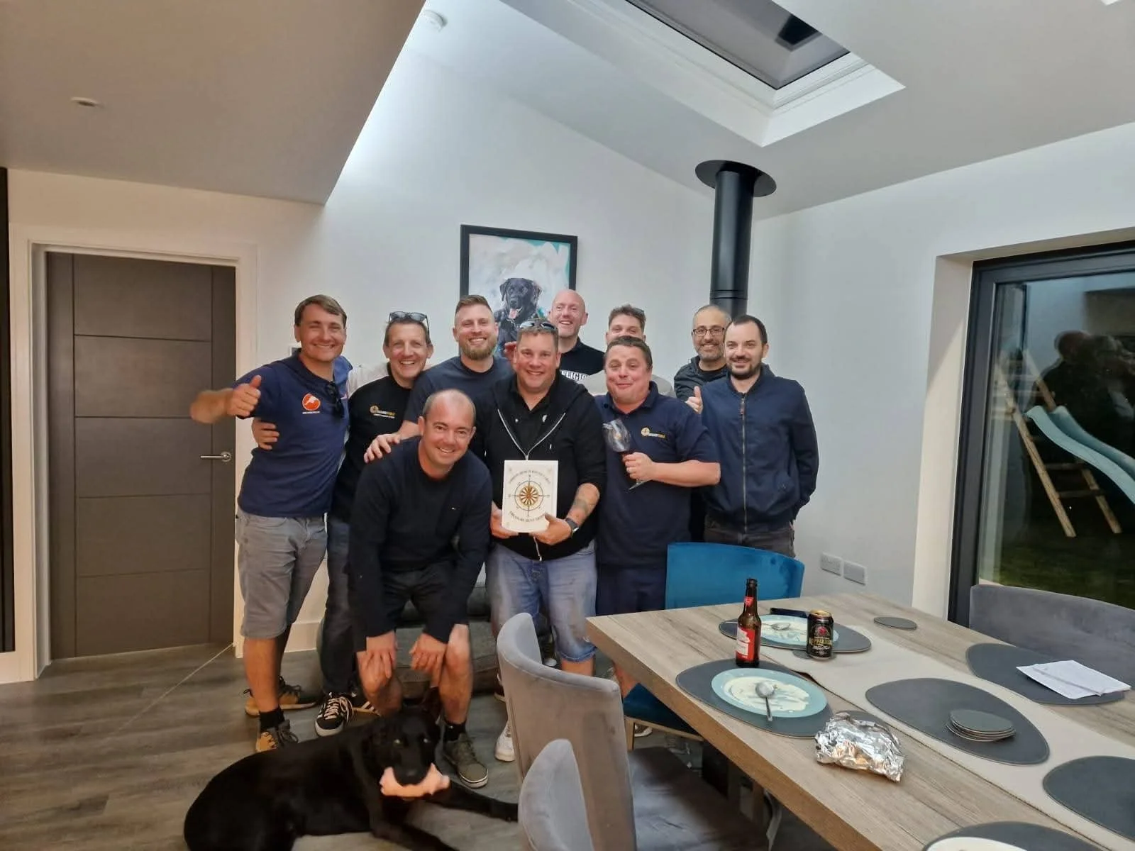 A group of nine men from Christchurch Round Table and a black dog gathered around a dining table in a modern room, smiling at the camera, with drinks and plates on the table, and a picture of a dog hanging on the wall behind them.