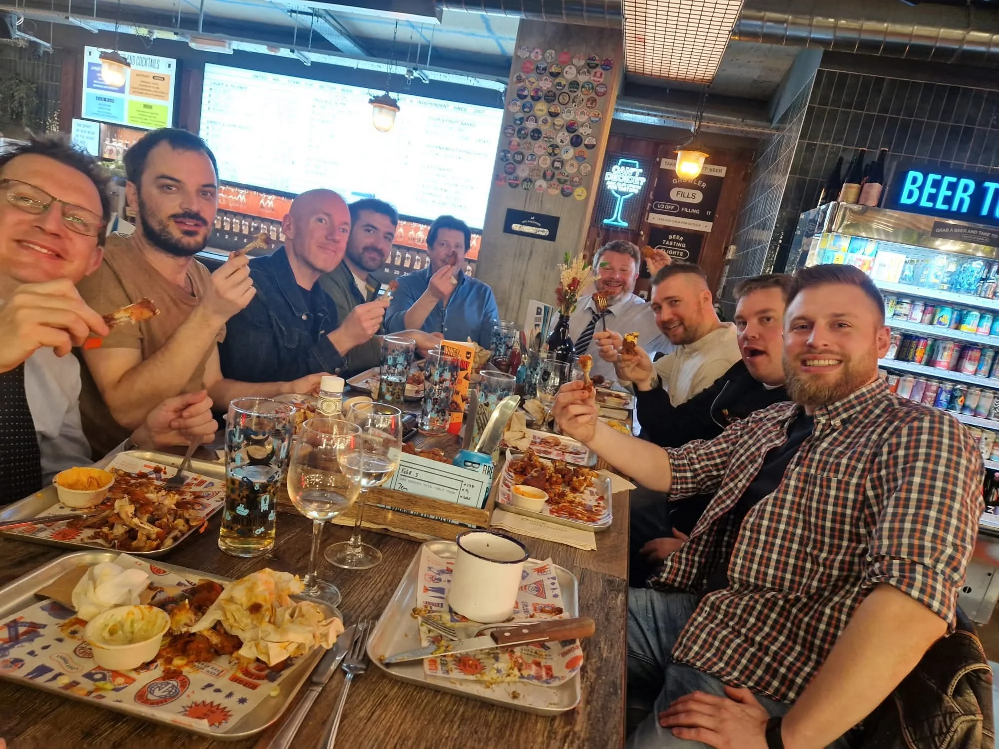A group of ten men sitting at a long table in a restaurant or bar, enjoying pizza and drinks. They are smiling and holding pizza slices, with a busy background of beer bottles and a digital menu on the wall.
