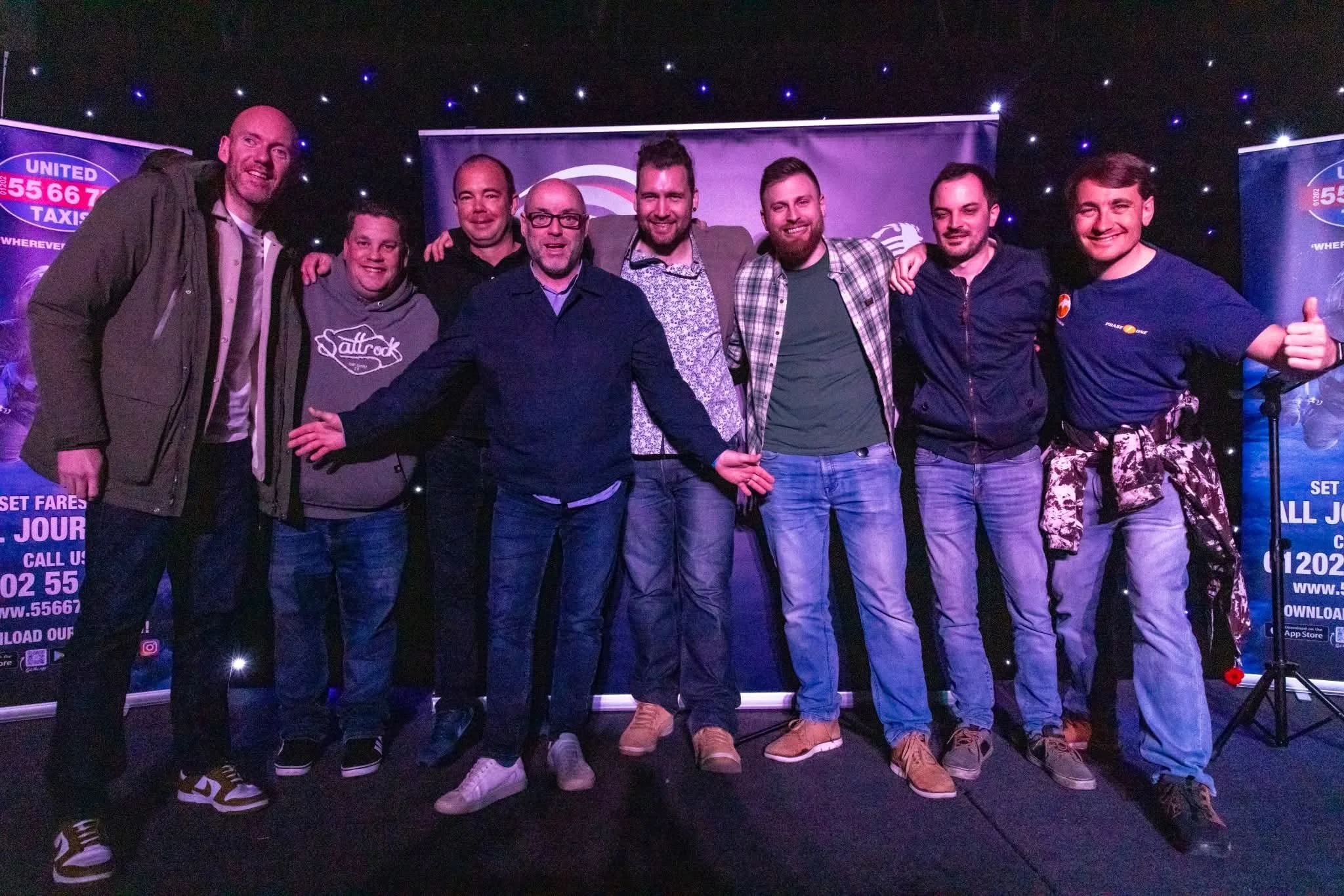 Group of nine men standing together on stage, smiling and posing for a photo with arms around each other, in front of a dark background with small lights and promotional banners.