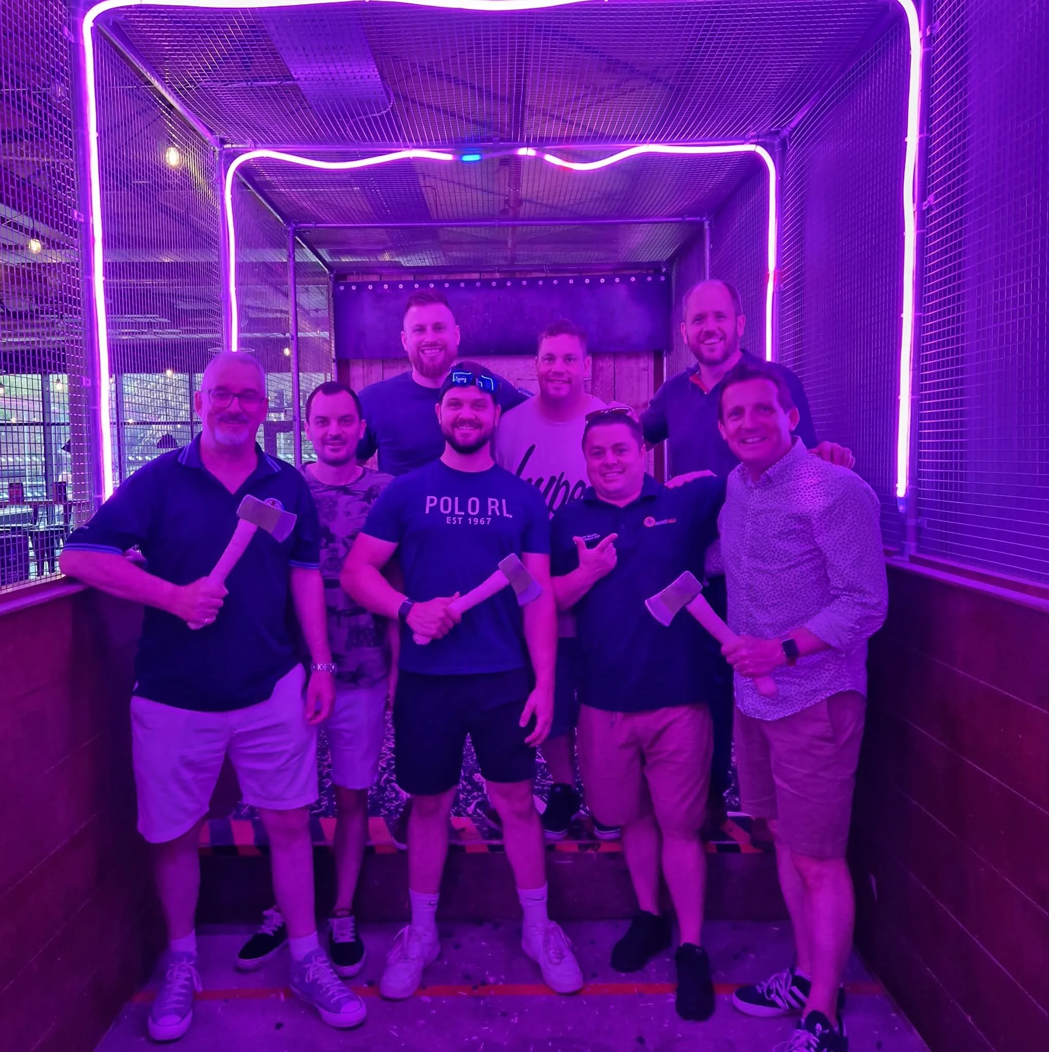 A group of nine smiling men from Christchurch Round Table holding axes inside a neon-lit, purple glow room, at an axe-throwing venue.