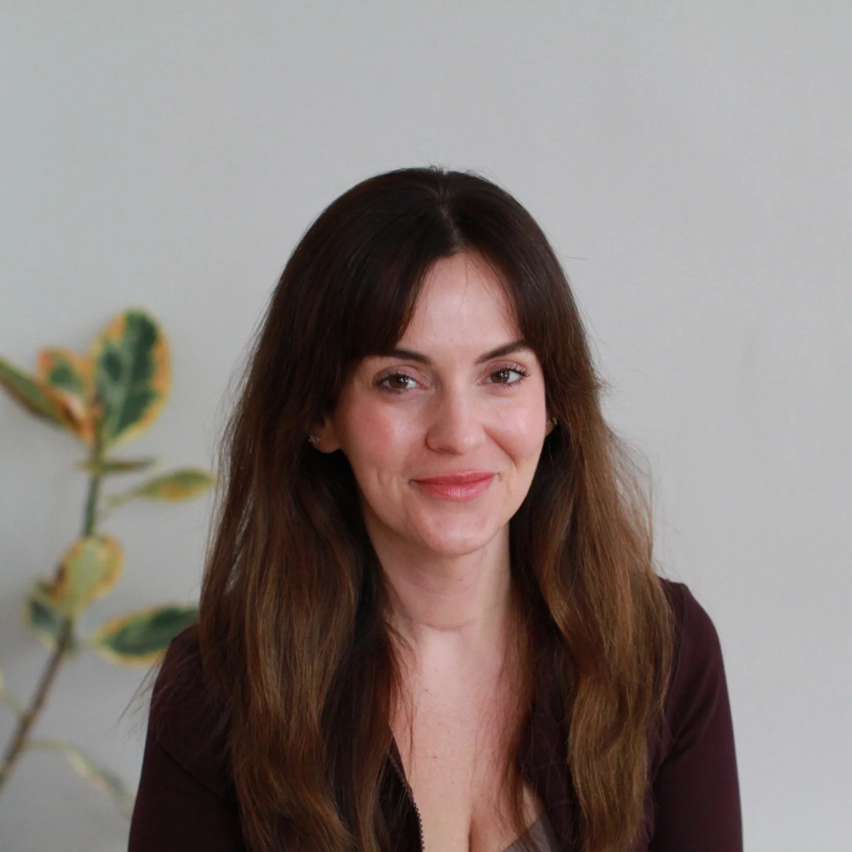 Portrait of a woman with long brown hair, wearing a dark top, smiling slightly, with a light-colored wall and a plant in the background.