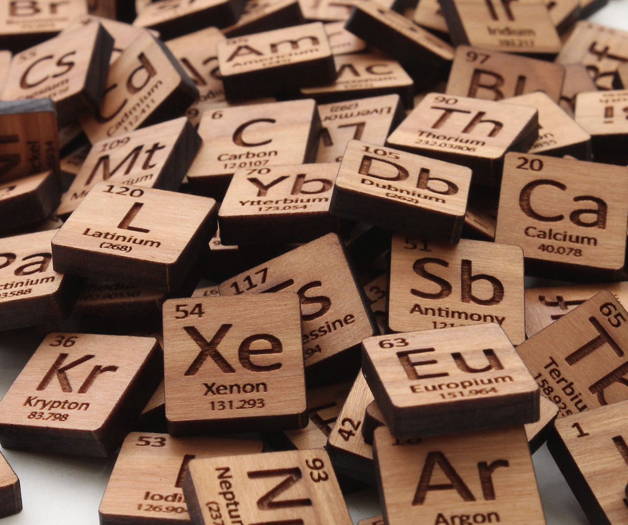 Close-up of scattered wooden Scrabble tiles featuring chemical element symbols and names, including Xenon, Chlorine, Barium, Calcium, and others.