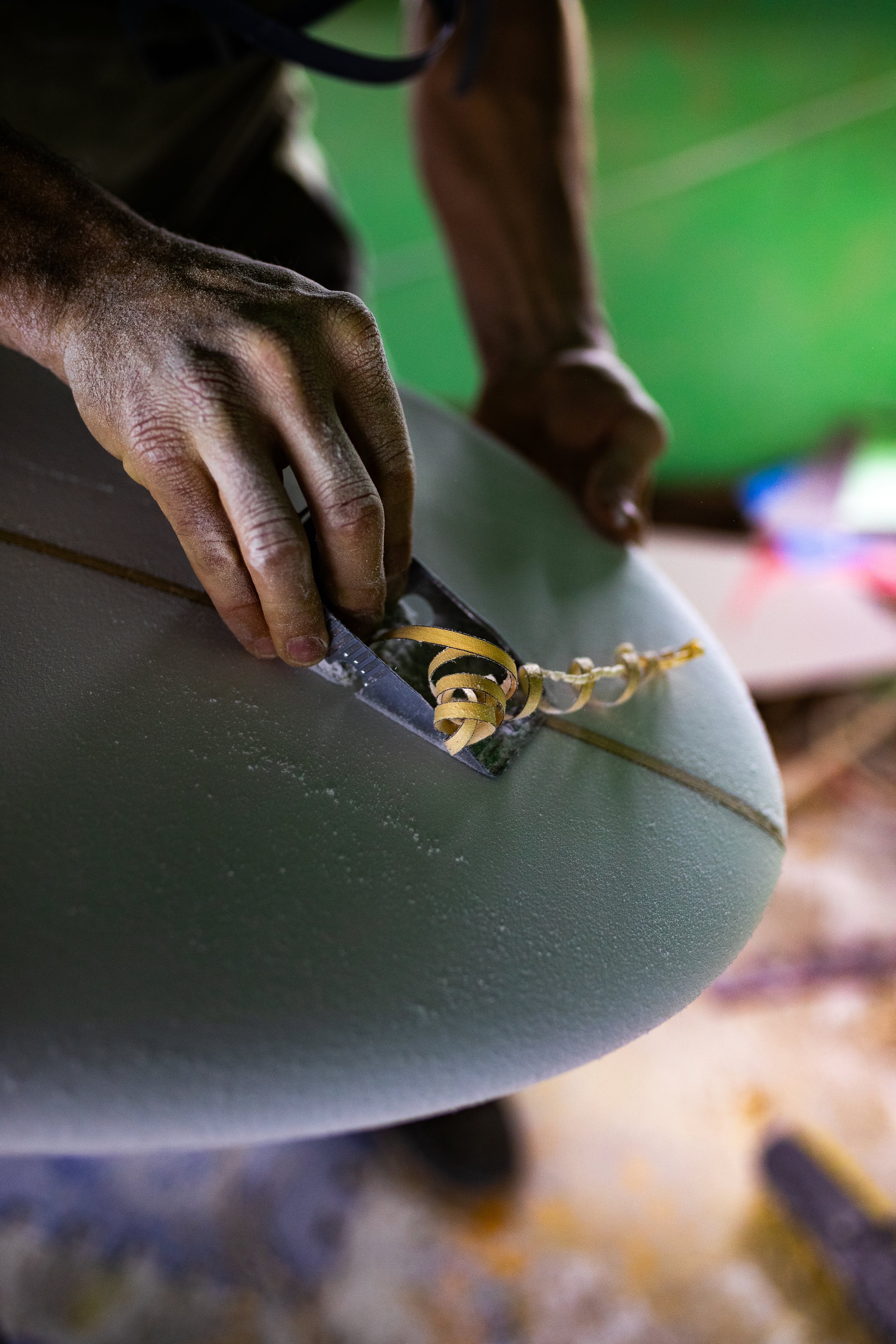 Person shaping a surfboard with a tool, creating wood shavings.