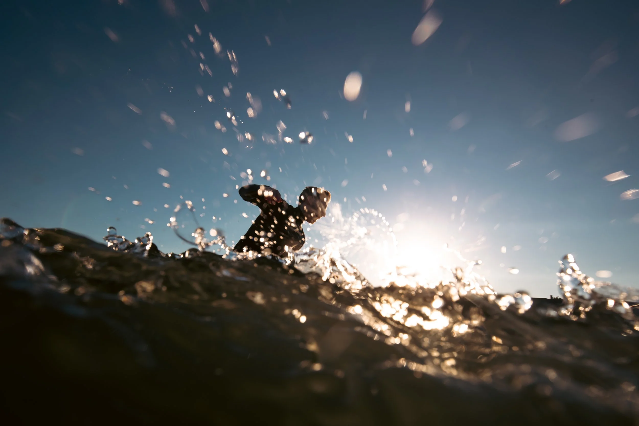 Silhouette of a person surfing in Maine, with the sun shining in the background and droplets in the air.
