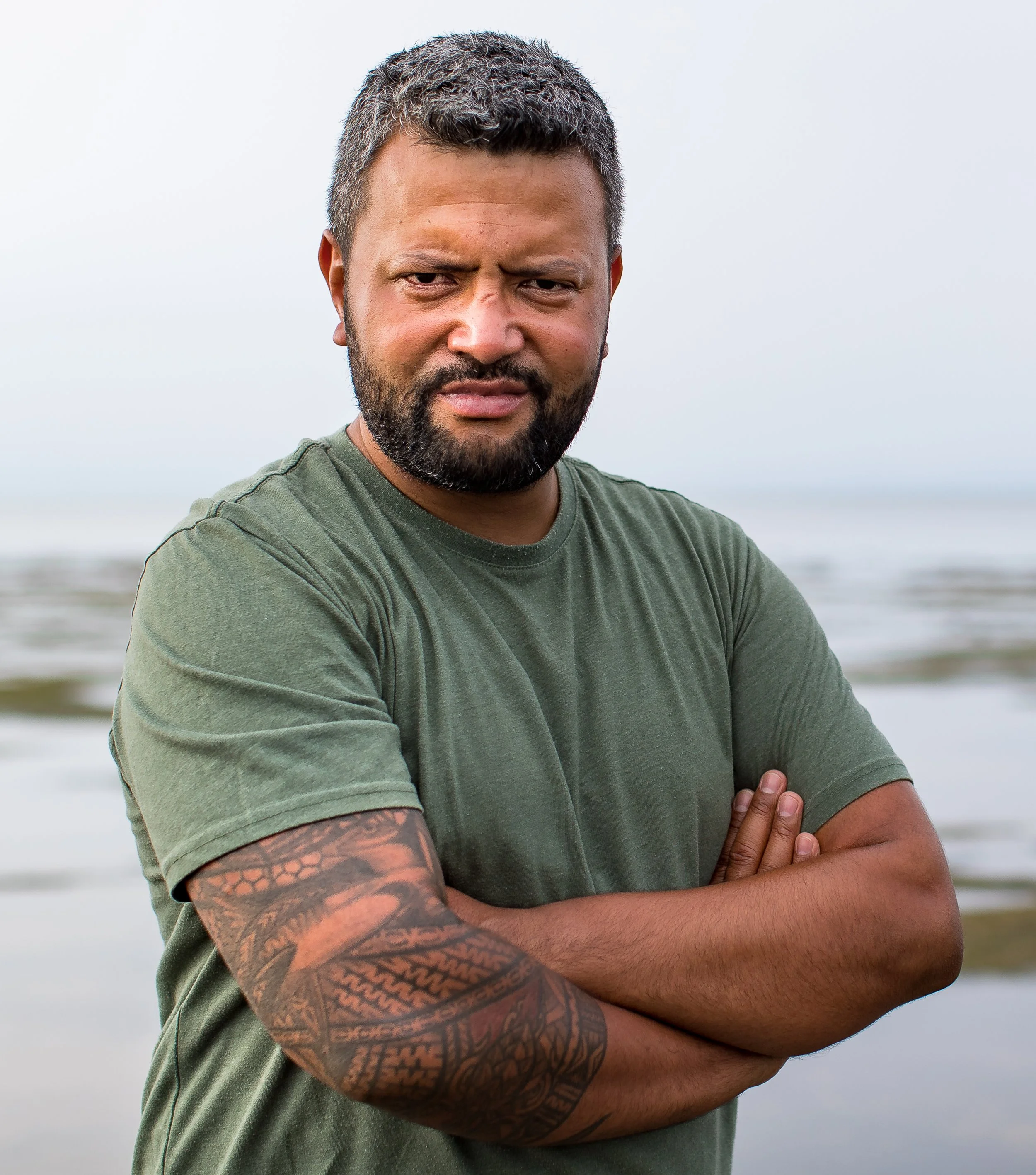 Man with short hair and beard wearing a green shirt, arms crossed, standing outdoors near water with a tattooed arm.