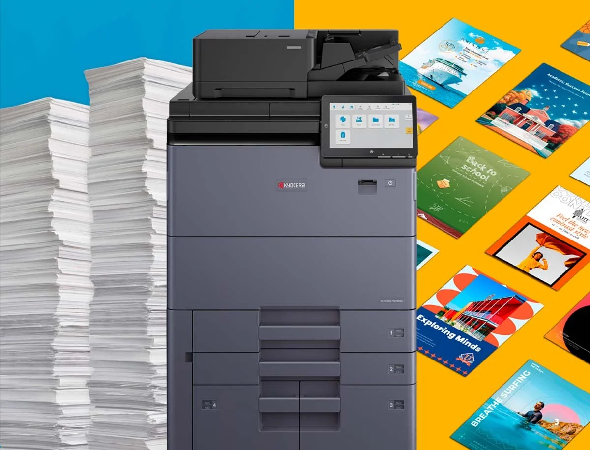 A modern black multifunction printer copier with a touchscreen interface, surrounded by large stacks of printed paper on the left, and various colourful educational and travel brochures on a yellow background on the right in Broken hill.