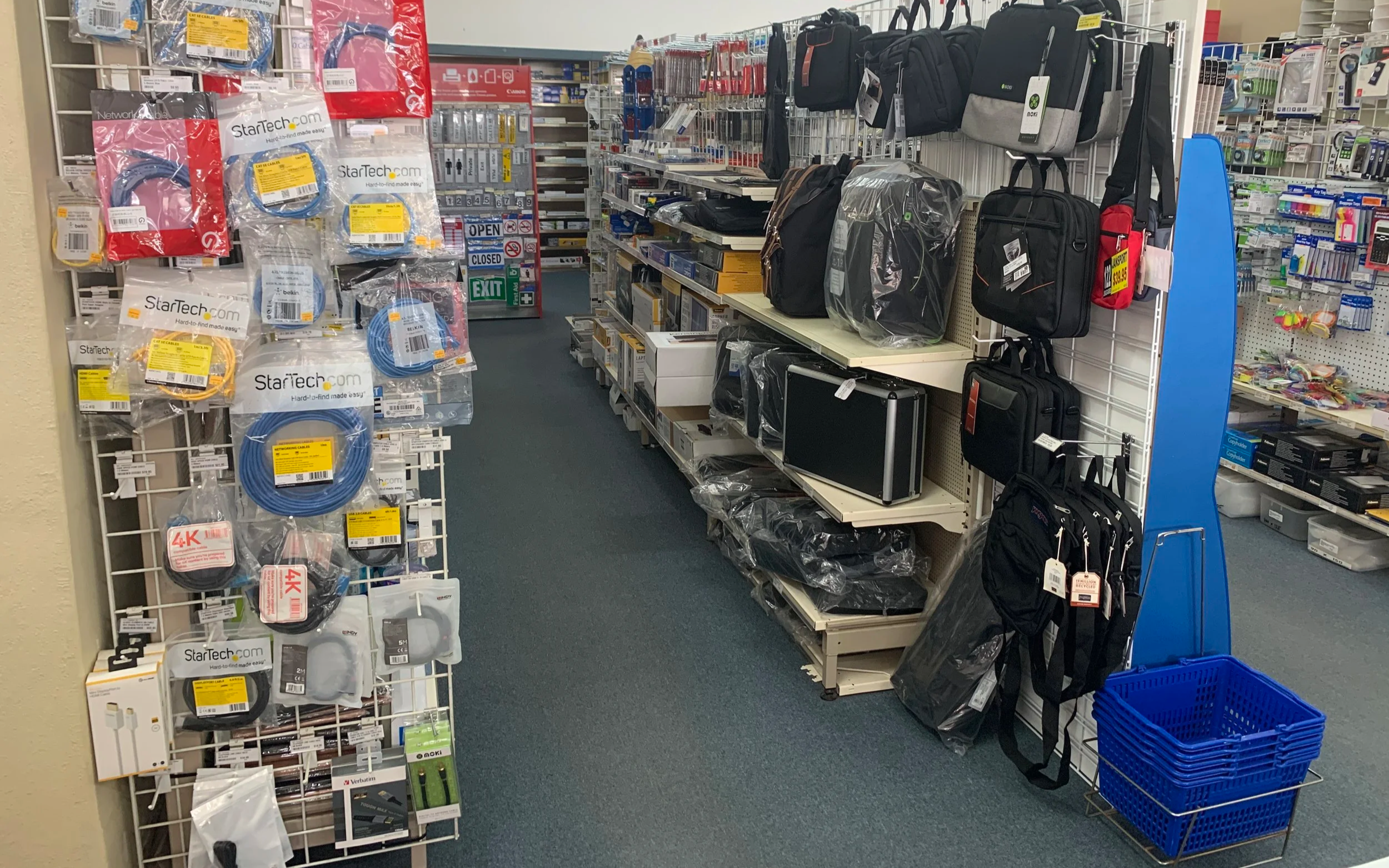 Aisle in an office supplies store with backpacks on the right and electronics accessories on the left, including cables and chargers. The store has a grey carpeted floor and a white grid wall display at Spicers Office Choice in Broken Hill.