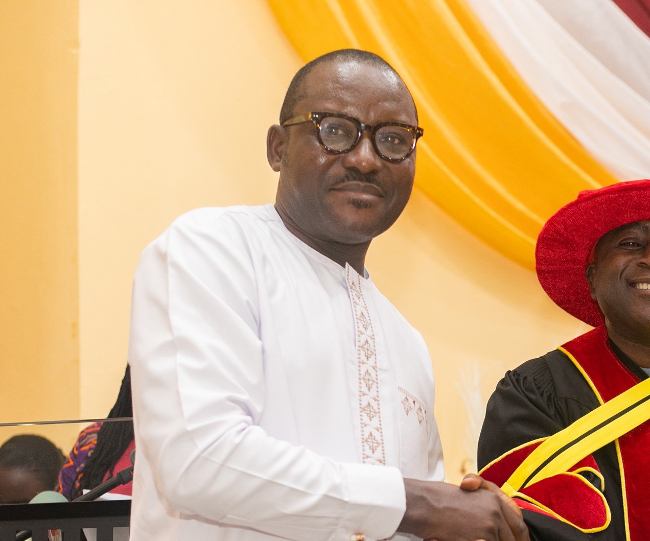 A man wearing glasses and a traditional white embroidered outfit standing next to a woman in ceremonial academic regalia with a red hat. They are at a formal event with a yellow and orange draped background.