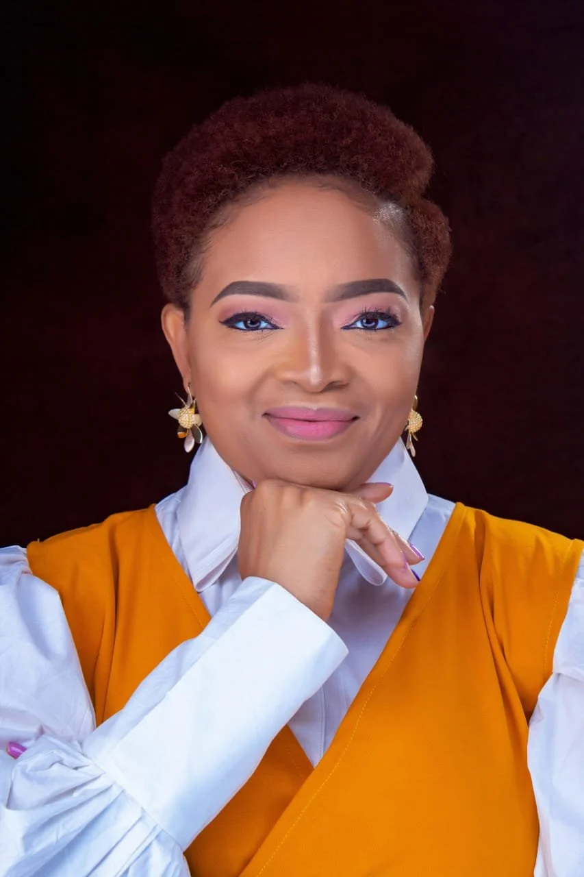 Portrait of an African woman with short curly hair, wearing makeup, earrings, a white shirt, and an orange vest, smiling with her hand under her chin against a dark background.