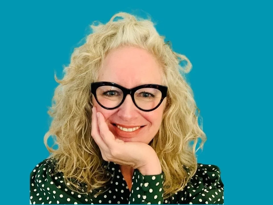 A woman with curly blonde hair wearing black glasses and a green blouse with white polka dots, smiling with her hand resting on her cheek against a blue background.