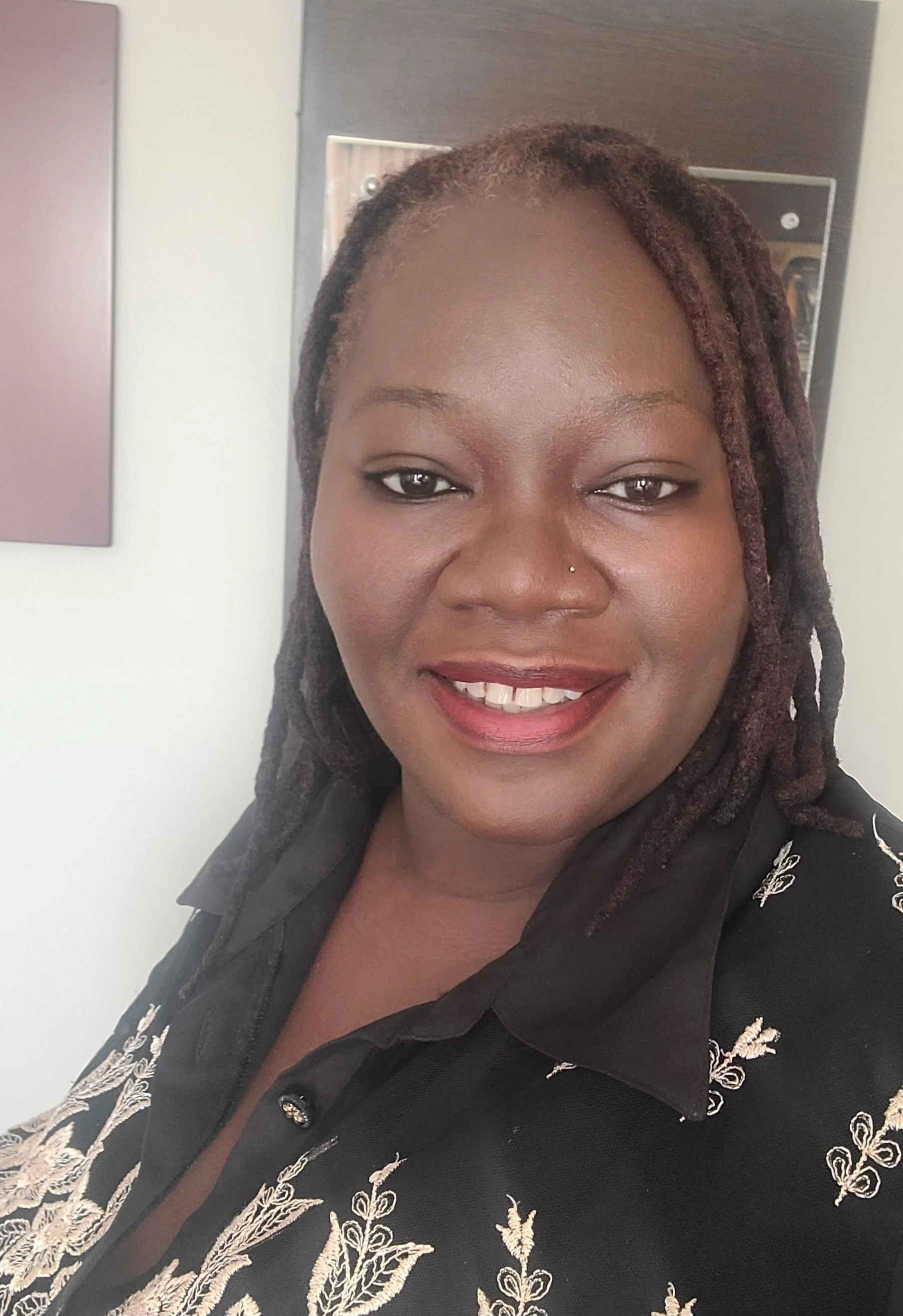 Close-up photo of a woman with dark skin, smiling, with shoulder-length dreadlocks, wearing a black top with embroidery.
