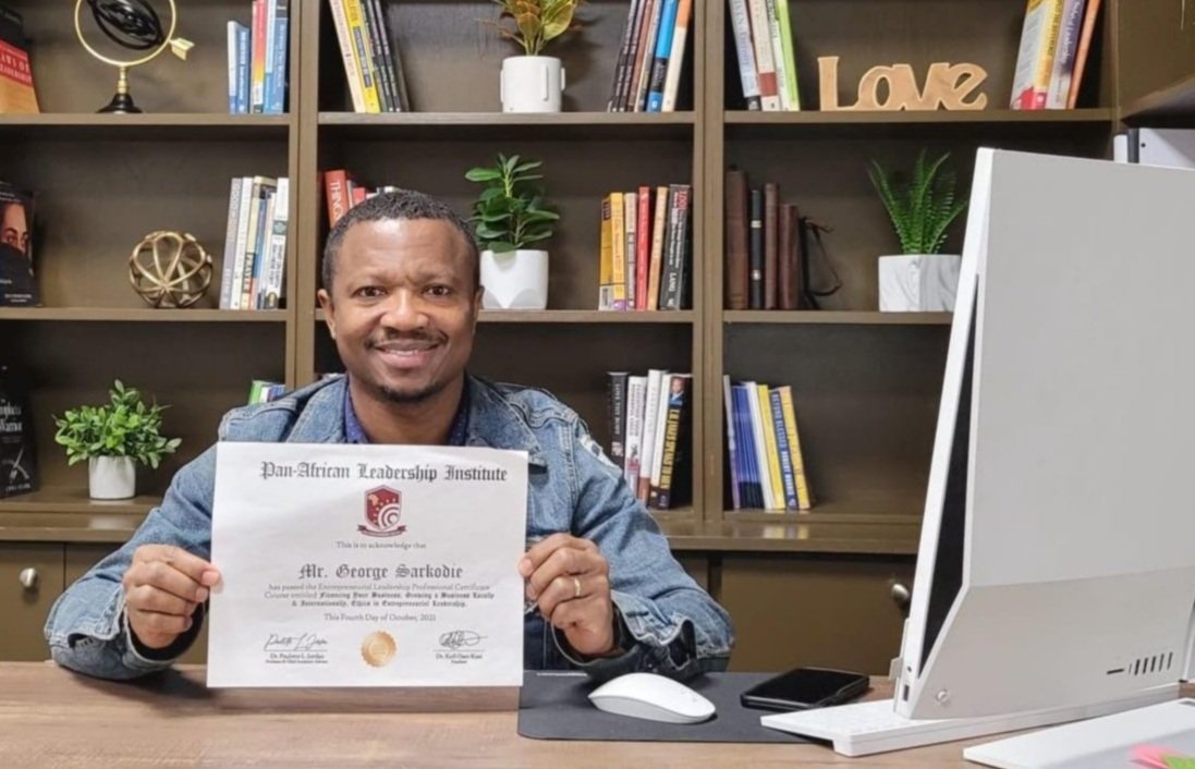 Man sitting at desk holding a certificate from the Pan-African Leadership Institute, smiling, with bookshelves and plants in the background.