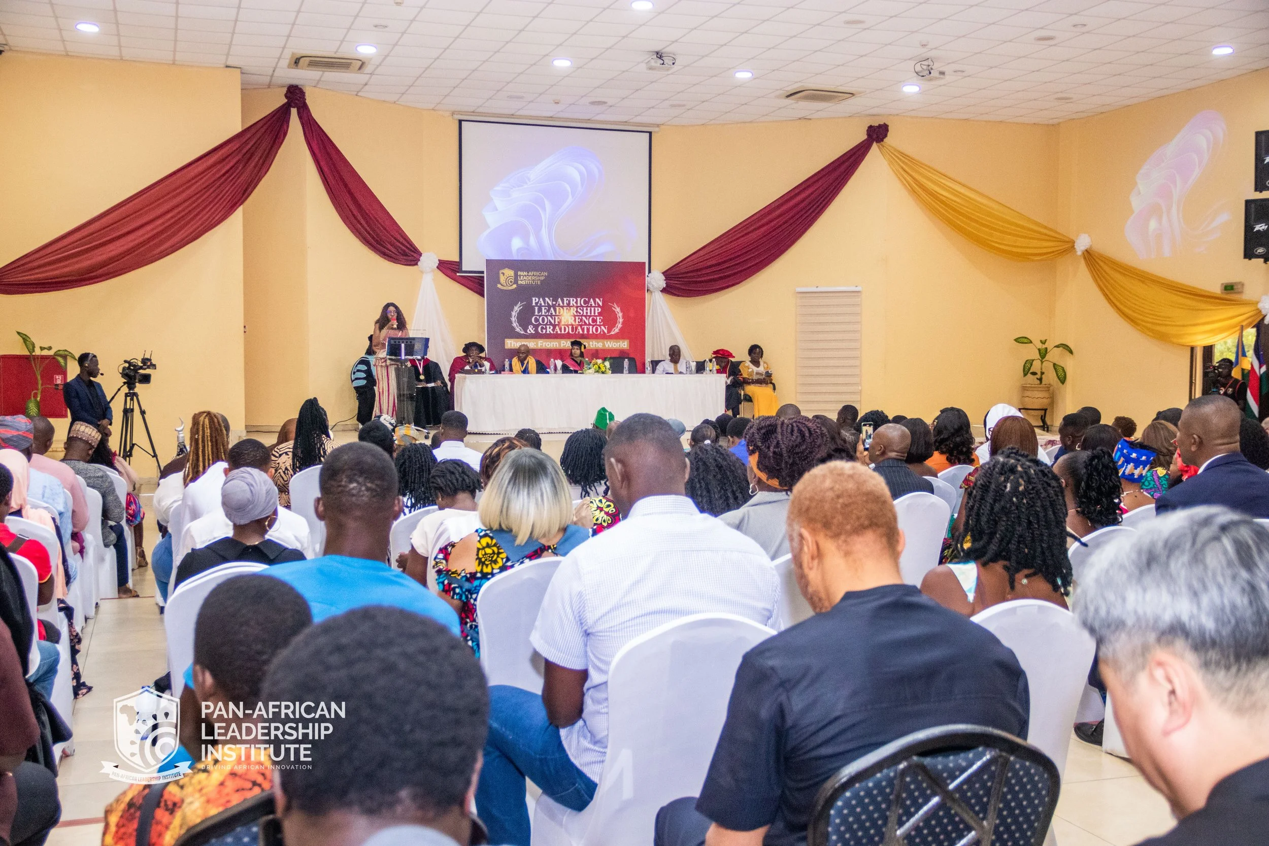 A large conference room filled with people attending the Pan-African Leadership Conference and Graduation. There is a stage with a banner, a panel of speakers, and decorations with drapes on the walls.
