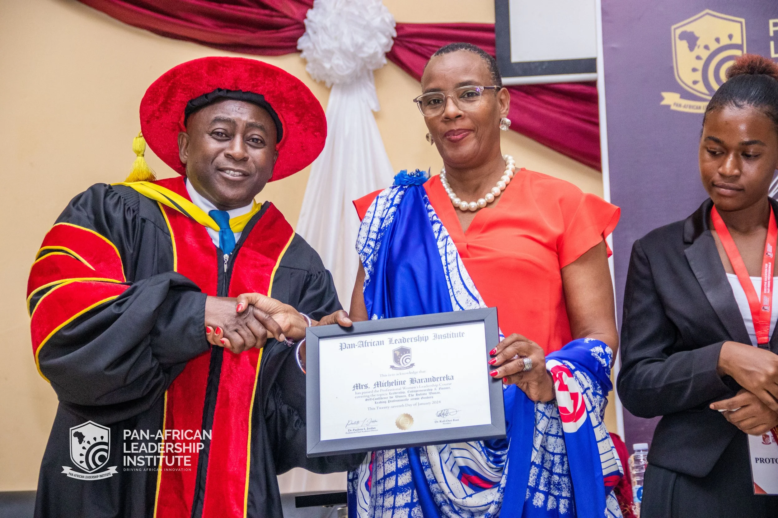 A man in academic regalia and a woman in a blue and orange dress shake hands and smile while holding a framed certificate during an awards ceremony. There is a woman to the right looking down and a banner with the logo of the Pan-African Leadership I