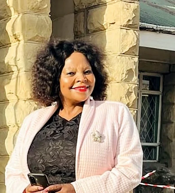 Woman with curly hair smiling, wearing a pink jacket over a black top, standing outside a building with stone wall and window.