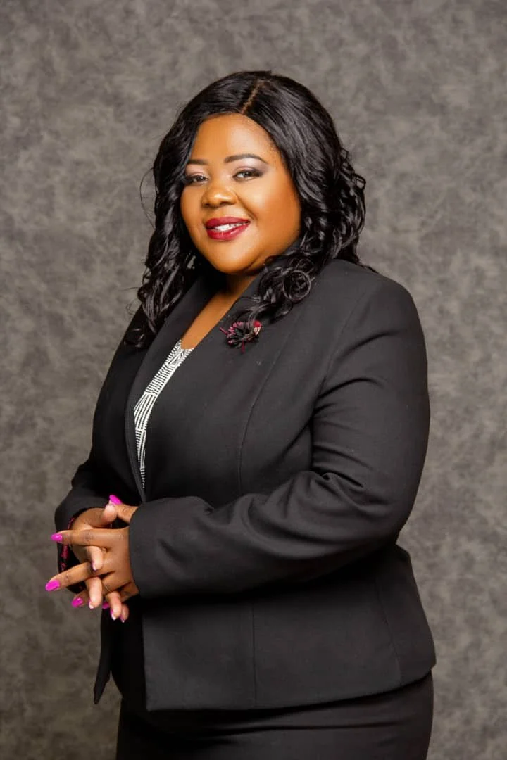A woman with black curly hair and red lipstick wearing a black blazer with a pin, standing against a gray textured background