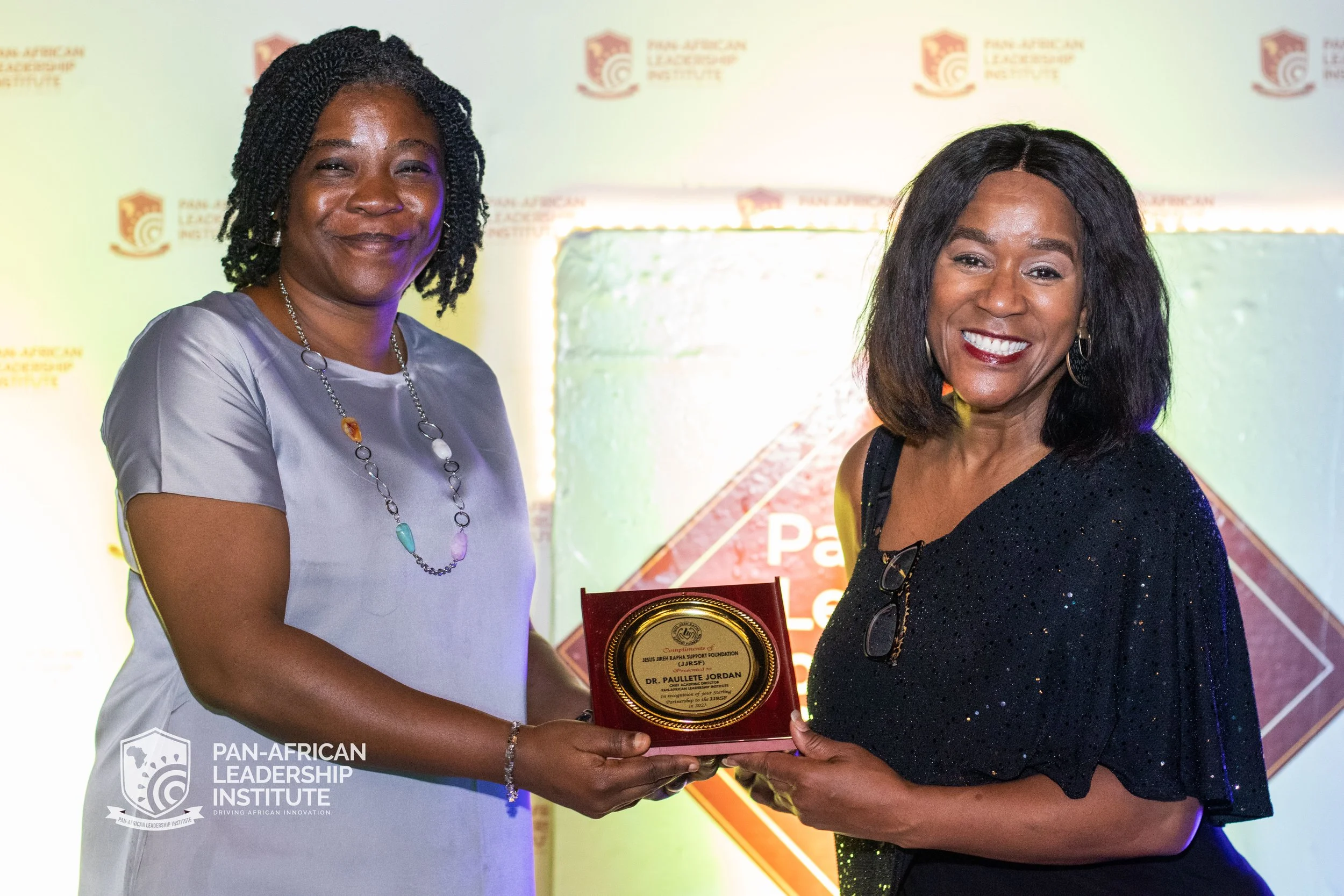 Two women are standing together at an award ceremony, one woman is giving an award plaque to the other. The background features the logo of the Pan-African Leadership Institute.