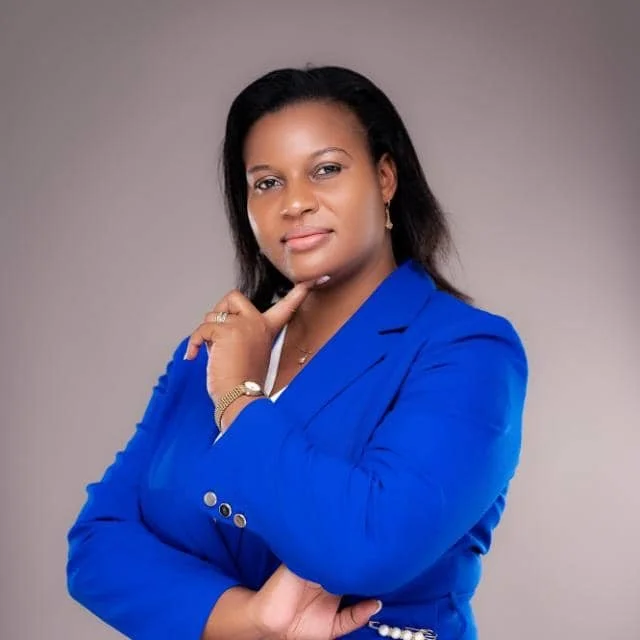 A woman with black hair, wearing a blue blazer, posing with her hand on her chin against a plain background.