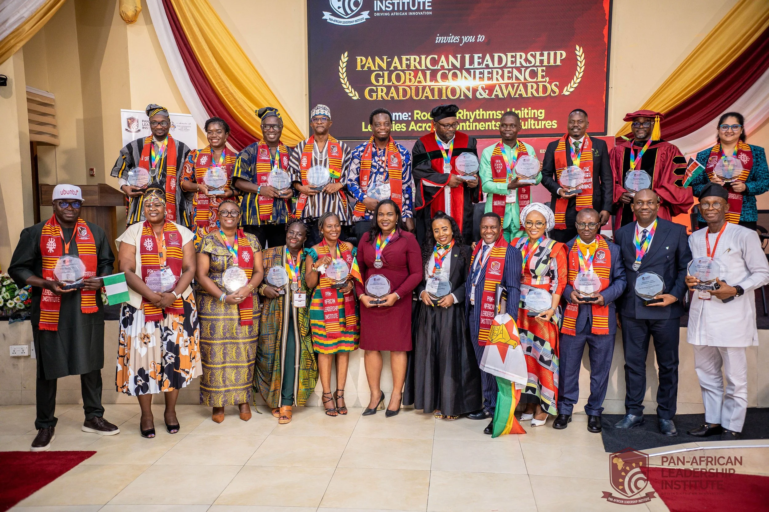 Group of people in formal attire, some wearing graduation robes and caps, standing on stage holding awards at the Pan-African Leadership Institute Conference, with a large screen displaying the event information behind them.