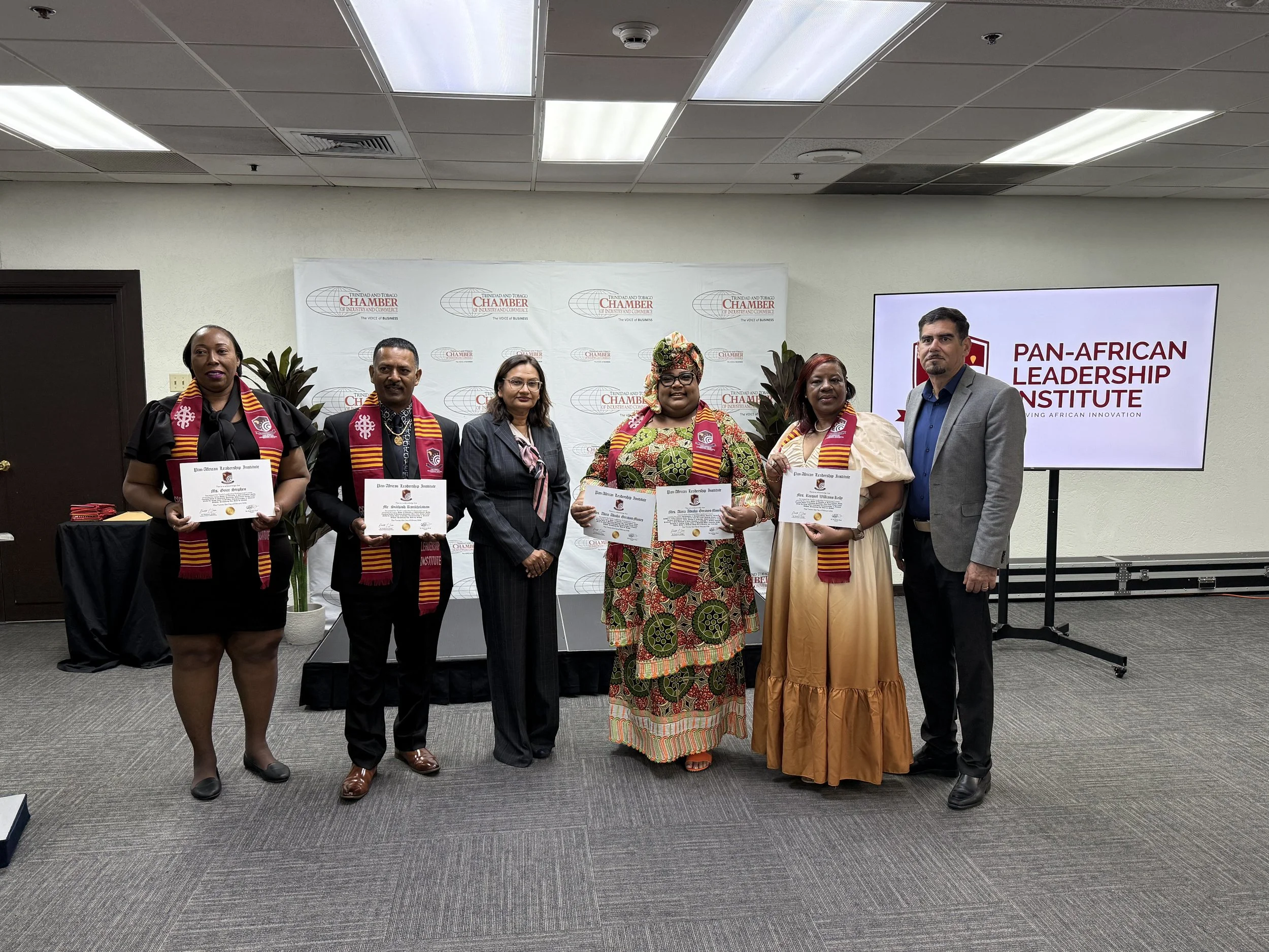 A group of seven people standing together in a conference room, holding certificates, with a banner of the Trinidad and Tobago Chamber of Industry and Commerce and a screen displaying 'Pan-African Leadership Institute' in the background.