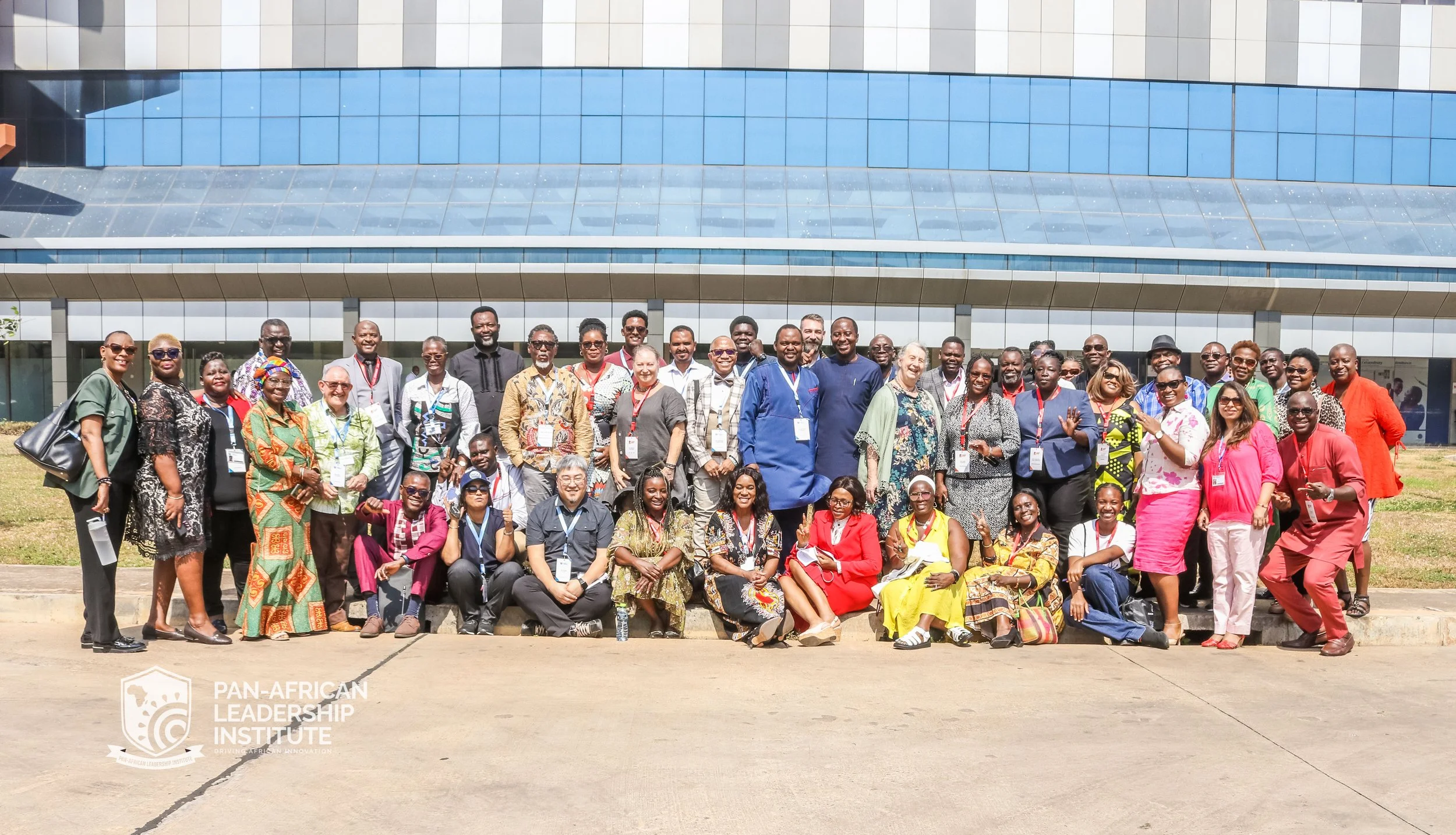 A large group of diverse people posing for a photo in front of a modern glass building during the day. Many are wearing conference badges around their necks, indicating they are attending an event related to the Pan-African Leadership Institute.