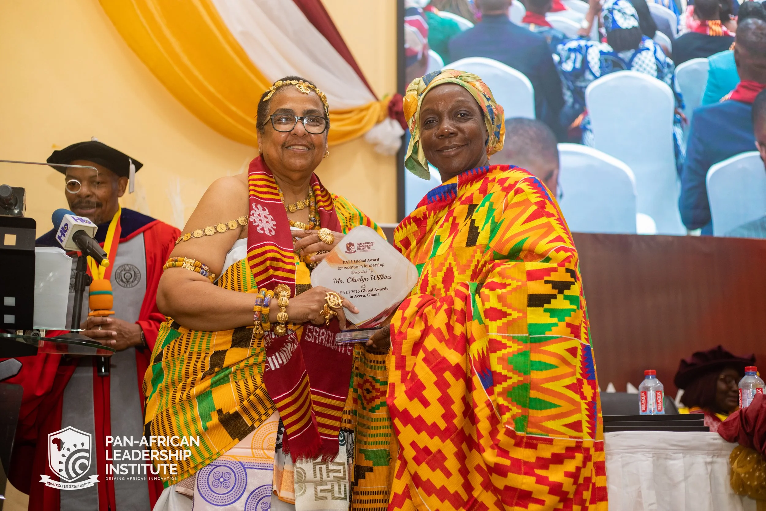 A woman wearing colorful traditional African attire receiving an award from another woman at the Pan-African Leadership Institute event. The scene is set in a decorated indoor venue with people seated in the background.