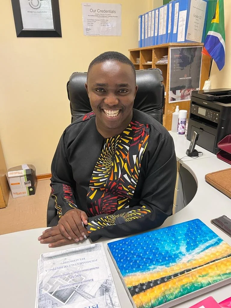 Smiling man sitting at an office desk with colorful folders and documents, South African flag and certificates on the wall.