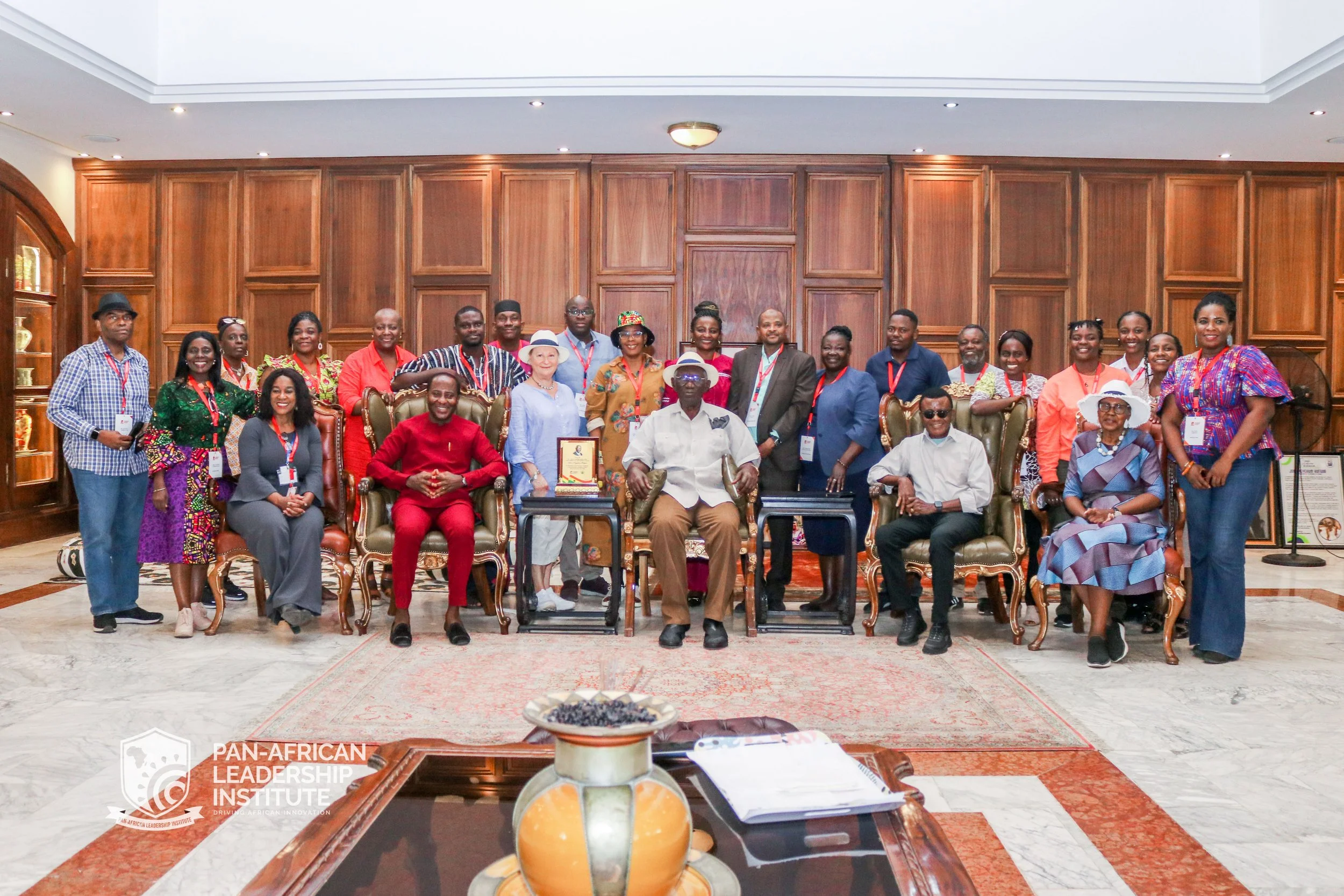 Group of 20 people, including men and women of African descent, gathered in a formal setting with a large wooden wall behind them. Some are seated in chairs, and others are standing, smiling for the photo. The setting appears to be an official or cel