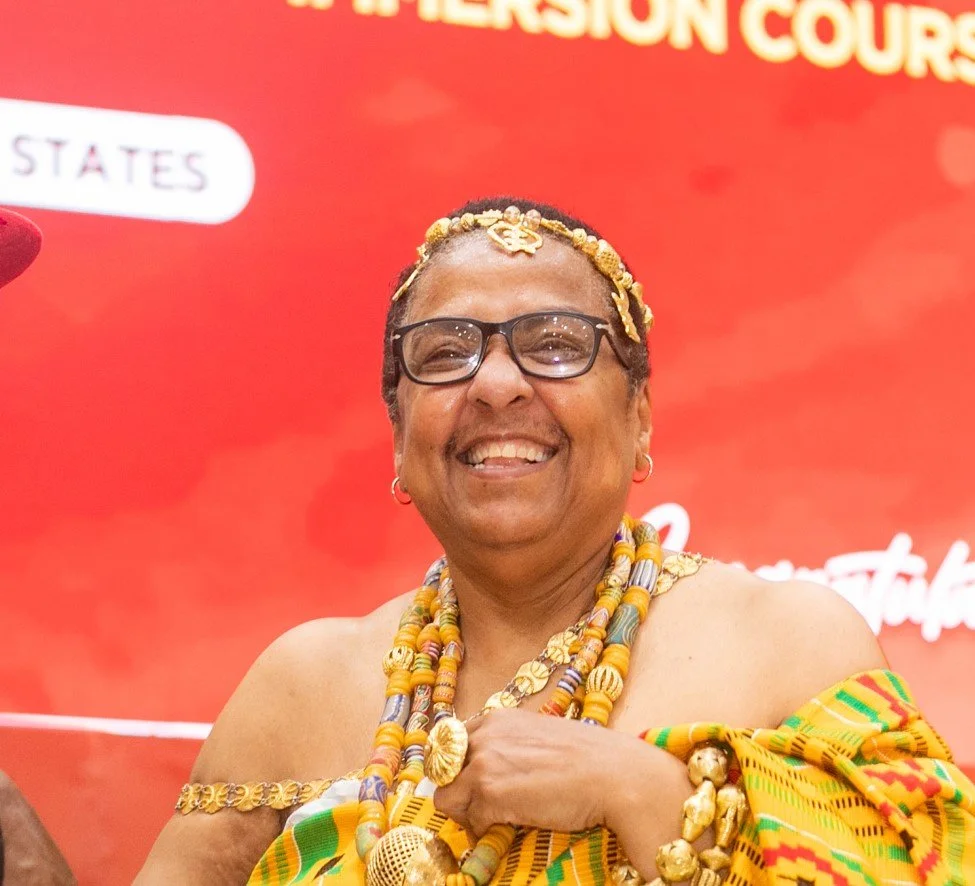A smiling woman in traditional African attire with gold jewelry and headpiece, against a red background.