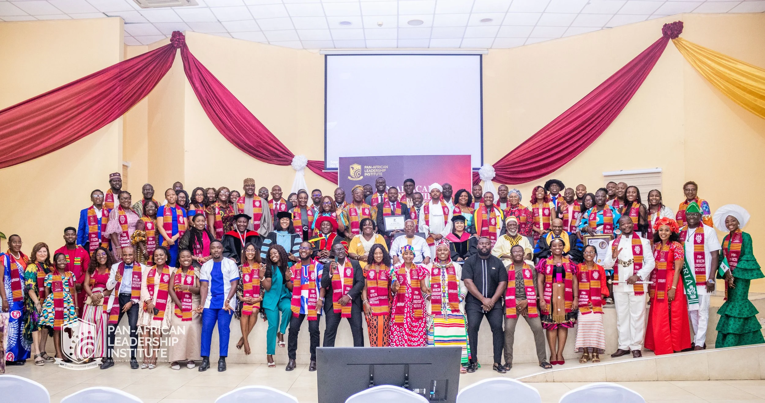 Group of men and women at a conference, some wearing traditional African attire and scarves with the logo of the Pan-African Leadership Institute. They are standing on stage in front of a yellow wall with red and gold drapes, and a large screen displ