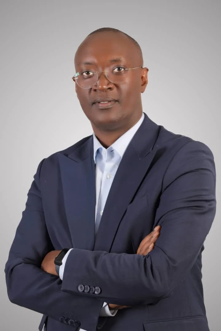 A man in a navy blue suit with a light blue shirt, wearing glasses, standing with arms crossed against a plain light grey background.