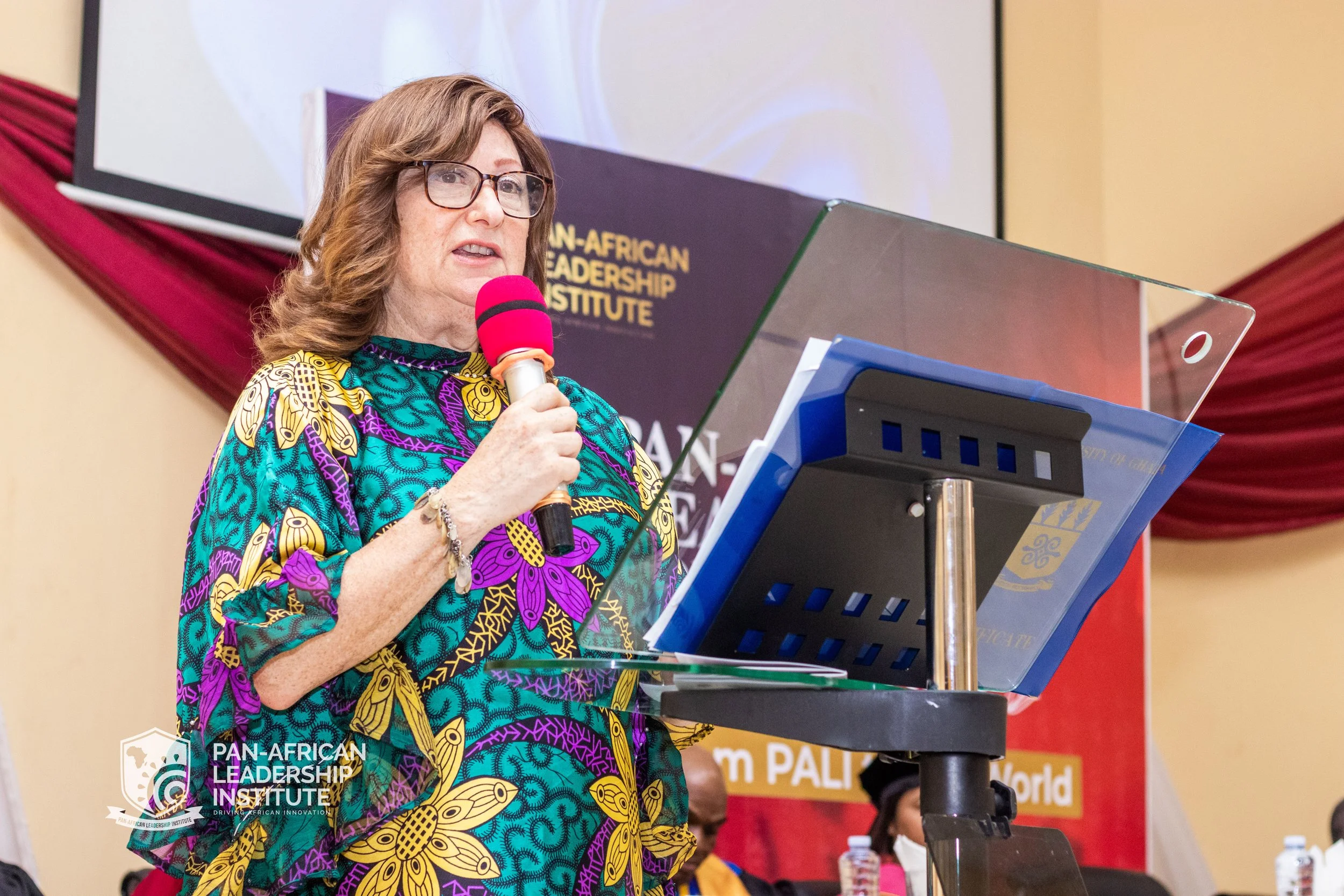 A woman with brown hair and glasses speaking at a podium, holding a pink microphone, wearing a colorful patterned dress with yellow and purple designs. Behind her is a sign that says 'Pan-African Leadership Institute'.