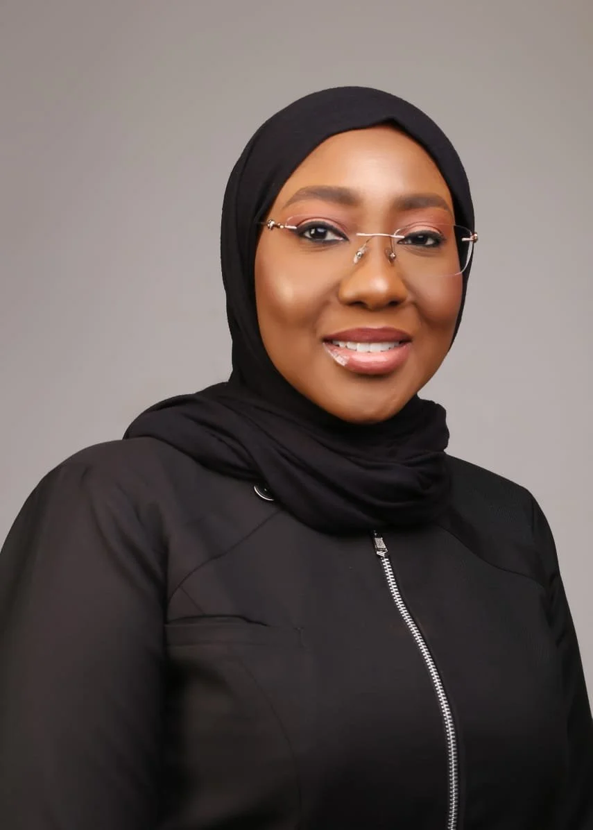 Portrait of a woman wearing a black hijab and glasses, smiling, with a plain gray background.