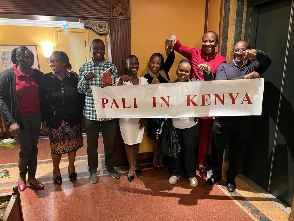 Group of eight people holding a banner that reads 'PALI IN KENYA' inside a room with wooden paneling and artwork on the walls, smiling and posing for the photo.