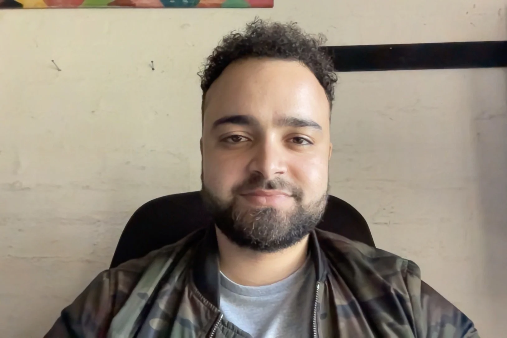 A man with curly hair and a beard, wearing a plaid jacket over a gray shirt, sitting in front of a cream-colored wall with small hooks and a black ledge.