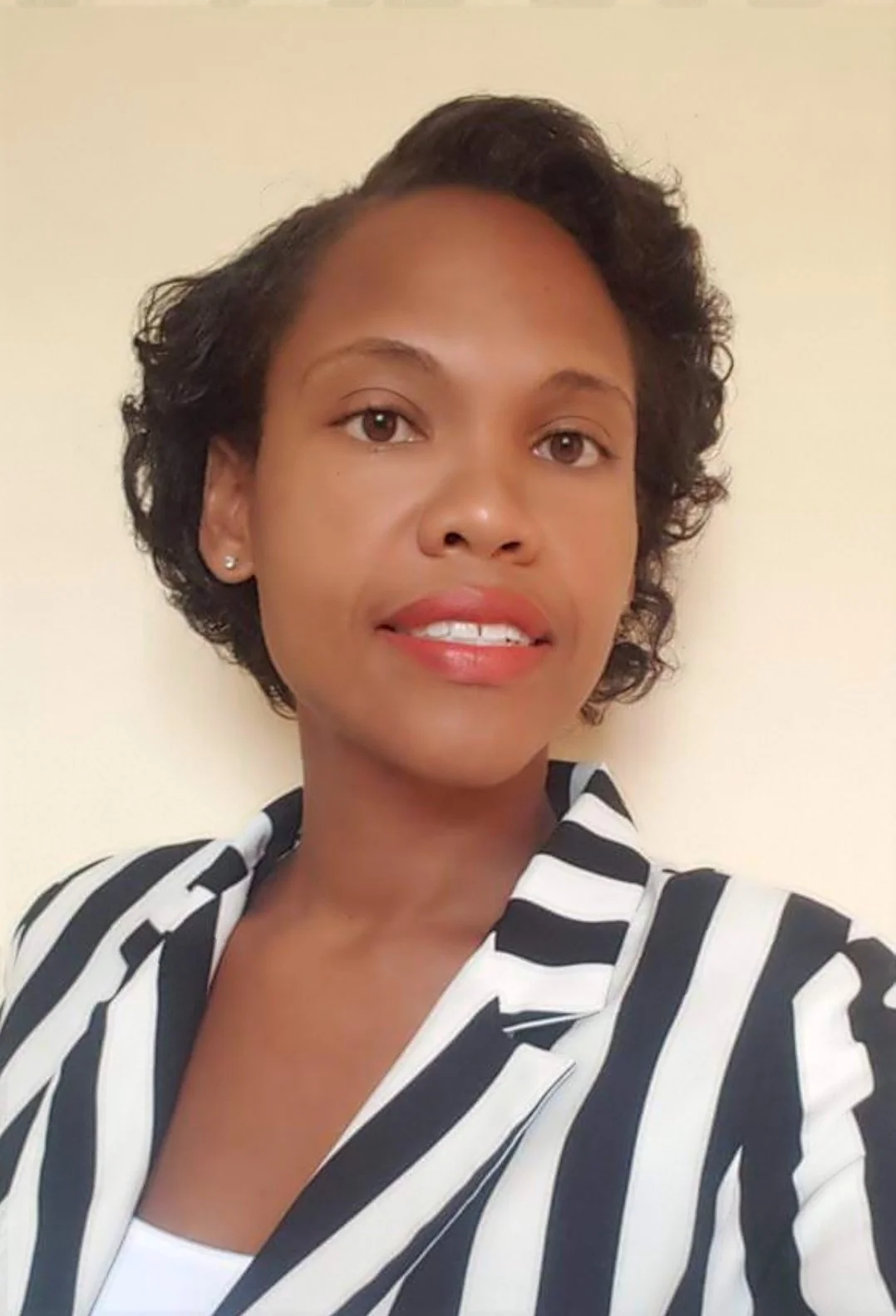 A woman with short, dark, curly hair wearing a black and white striped blazer and small earrings, standing against a plain wall.