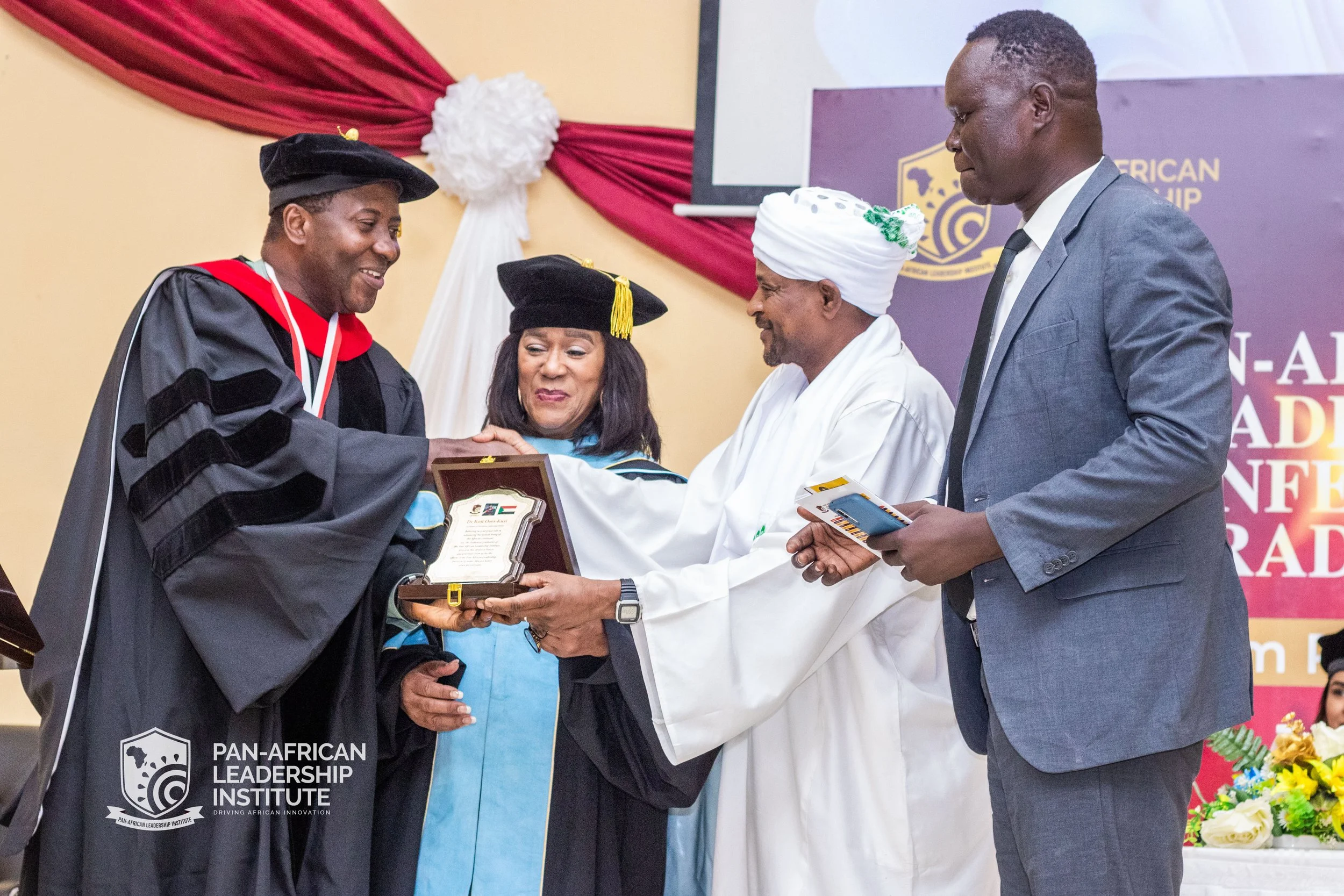 A man in academic regalia shaking hands with a woman holding a plaque. Two other men, one in traditional African attire and a gray suit, are observing at a ceremony with a banner for the Pan-African Leadership Institute in the background.