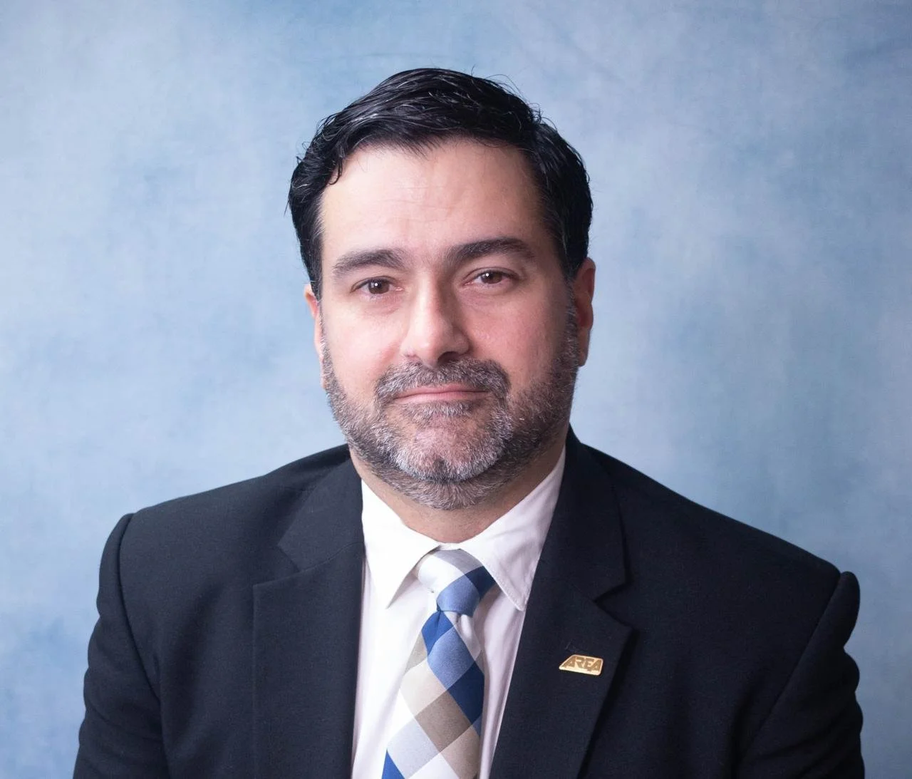 A professional man with dark hair and a beard, wearing a black suit, white shirt, and a striped grey, white, and blue tie, poses for a portrait against a light blue background.