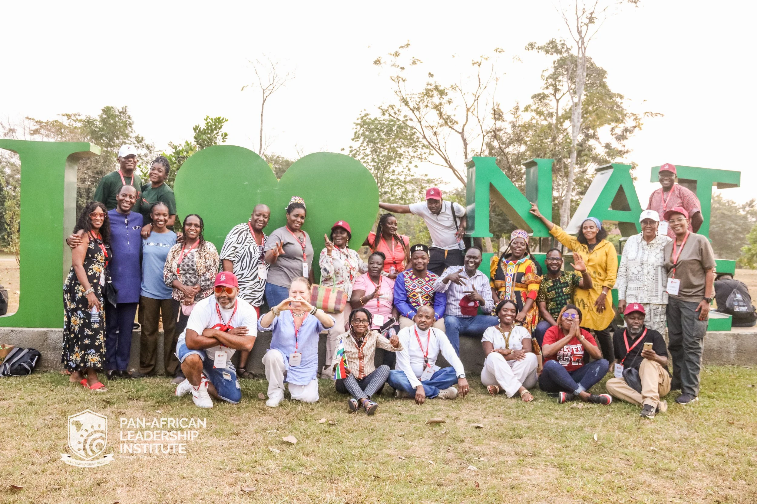 Group of diverse people gathered outdoors in front of large green 'I LOVE NAIROBI' sign on a grassy area with trees. Some are smiling and making gestures, wearing casual clothing and conference lanyards.