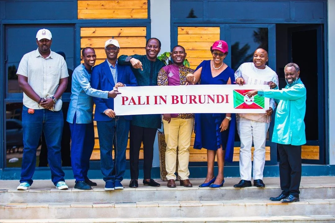 Group of nine people holding a banner that says 'PALI IN BURUNDI' and a Burundian flag, standing outside a building with blue walls and wooden accents.