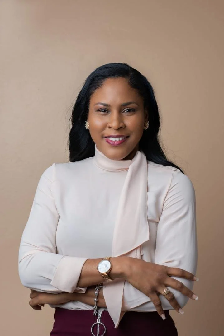 A woman with dark hair wearing a cream-colored blouse with a bow on the neck, gold earrings, a watch, and a ring, smiling and standing with arms crossed in front of a beige background.
