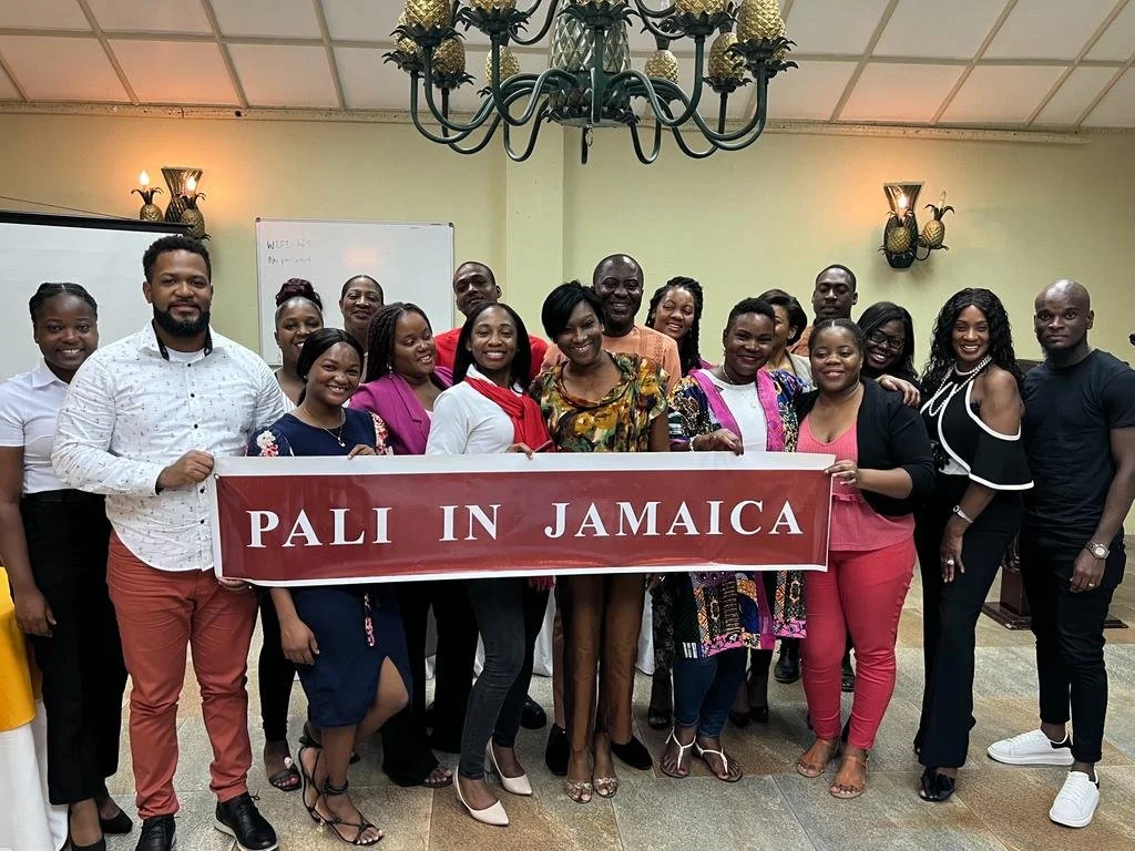Group of people holding a red banner that reads 'PALI IN JAMAICA,' standing in a room with a chandelier, whiteboard, and wall lights.