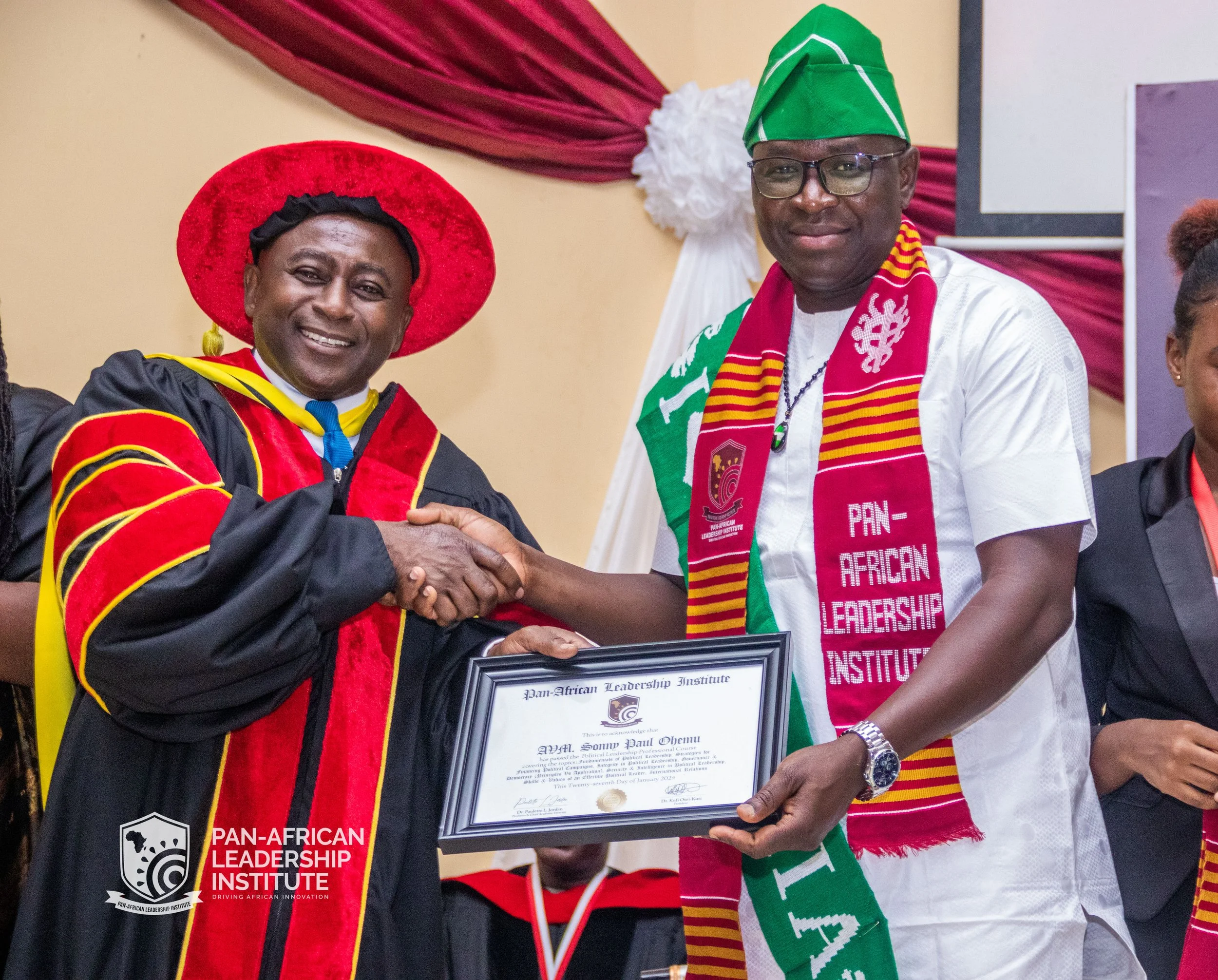 Two men at a formal event shaking hands, one of them wearing an academic cap and gown with colors of red and black, and the other dressed in a white shirt with a green scarf and a green cap, holding a framed certificate from the Pan-African Leadershi