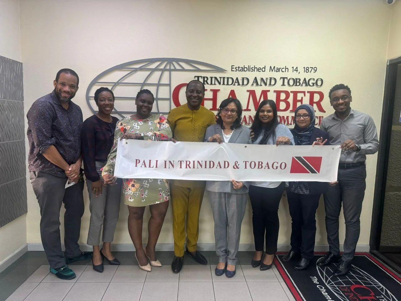 Group of nine diverse people holding a banner that reads "PALI IN TRINIDAD & TOBAGO" in front of a wall with the Trinidad and Tobago Chamber of Commerce logo.