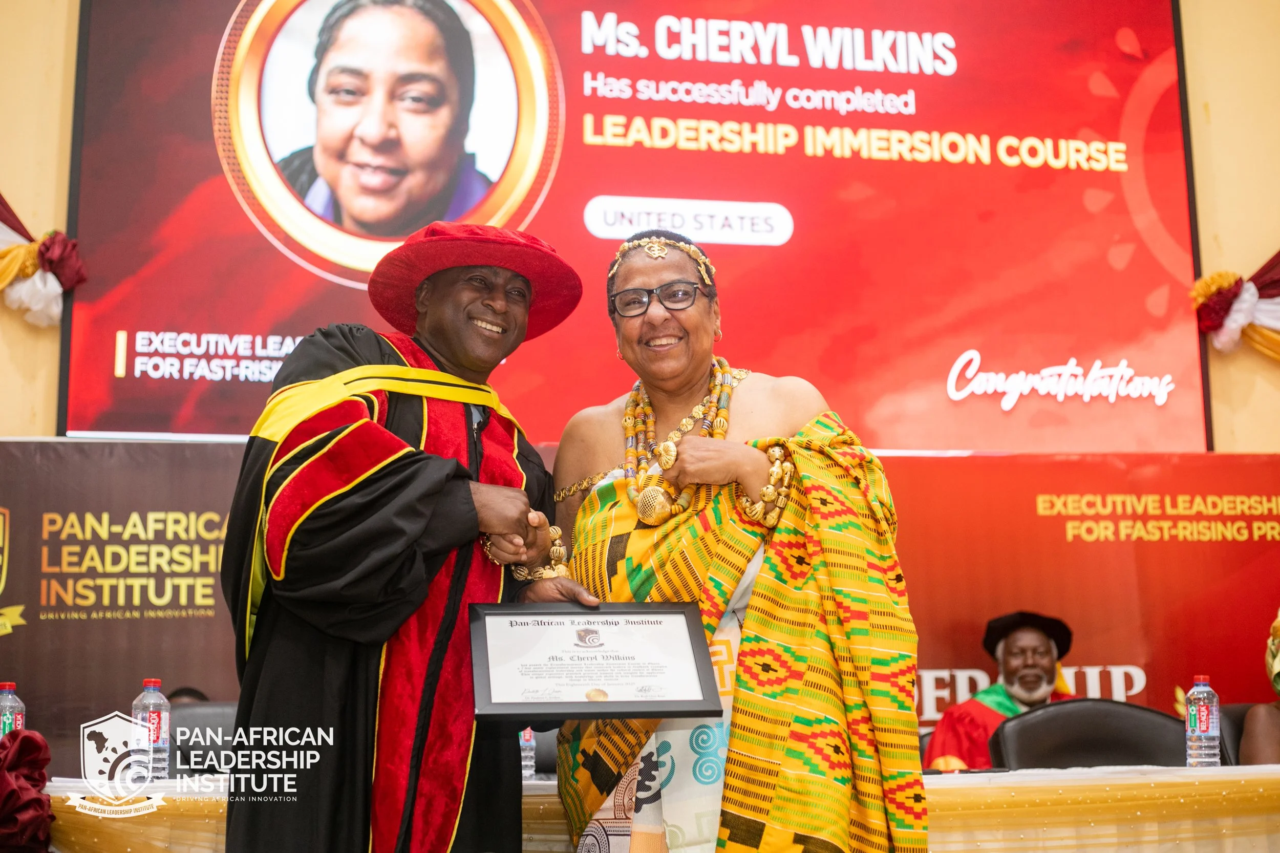 Two African women smiling and shaking hands at an award ceremony. One woman is dressed in traditional African attire with a yellow, green, and orange patterned cloth, and the other is in academic regalia with a red and black gown and a large red hat.