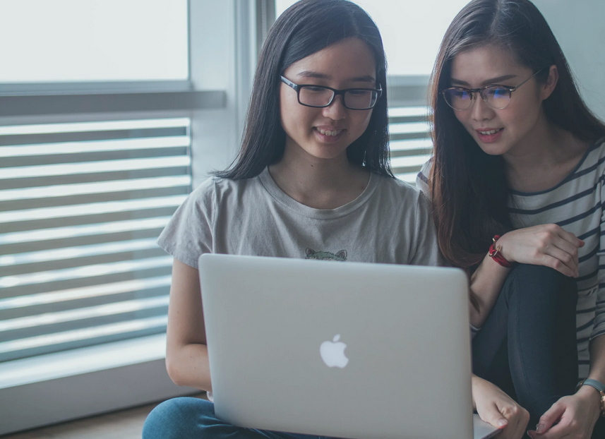 Two young women with glasses looking at a laptop together in a bright room.