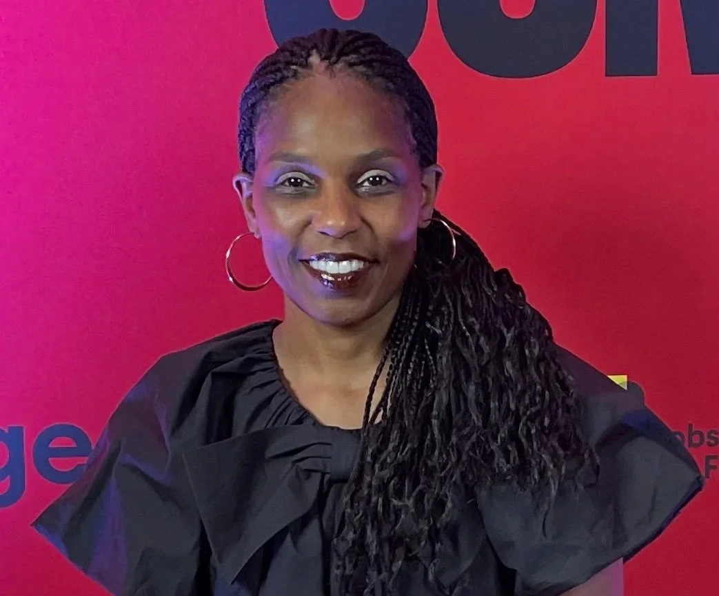 A woman with long braided hair wearing hoop earrings and a black top, standing in front of a pink background with black text.