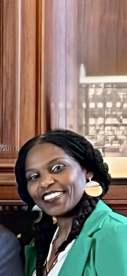 Smiling woman with braided hair wearing gold earrings and a green blazer inside a wooden-paneled room.